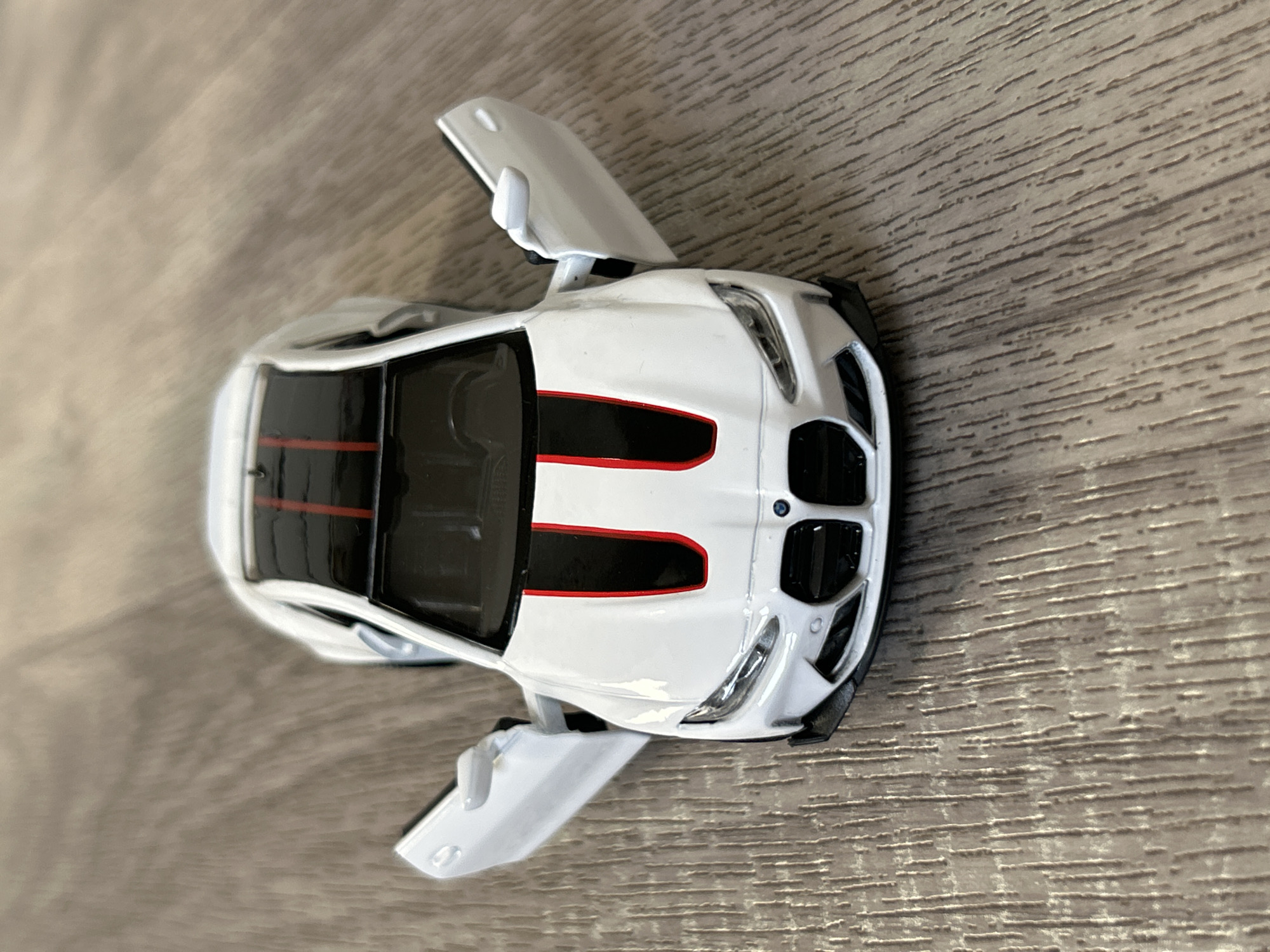 The image shows a white toy car on a wood-grain surface. The car has two doors open and red detailing on the black roof. The front of the car is visible, with a BMW logo on the hood. The image appears to be taken from directly above the car. There are no people visible.
The location of the city of Strasbourg, France, cannot be determined.