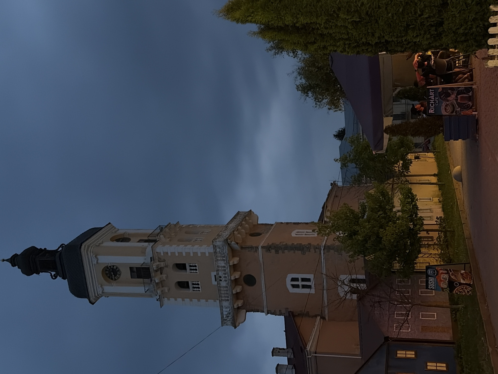 The image depicts the historic Town Hall Tower in Kamianets-Podilskyi, Ukraine, at dusk. The tower, a prominent architectural landmark, features a light-colored facade with decorative elements and a clock face visible on one side. To the left of the tower, a pastel-colored building with small windows is present. In the foreground, a sidewalk runs alongside a street, with a green lawn bordering it. A tall, dark green coniferous tree is situated on the right side of the frame. Beneath a large, dark blue awning, a café is set up with several patrons seated at tables. A sign for "RICHART CAFE" is visible in front of the café. Another sign advertising "SUSHI" stands to the left of the café. The overall atmosphere is calm and illuminated by the ambient twilight.