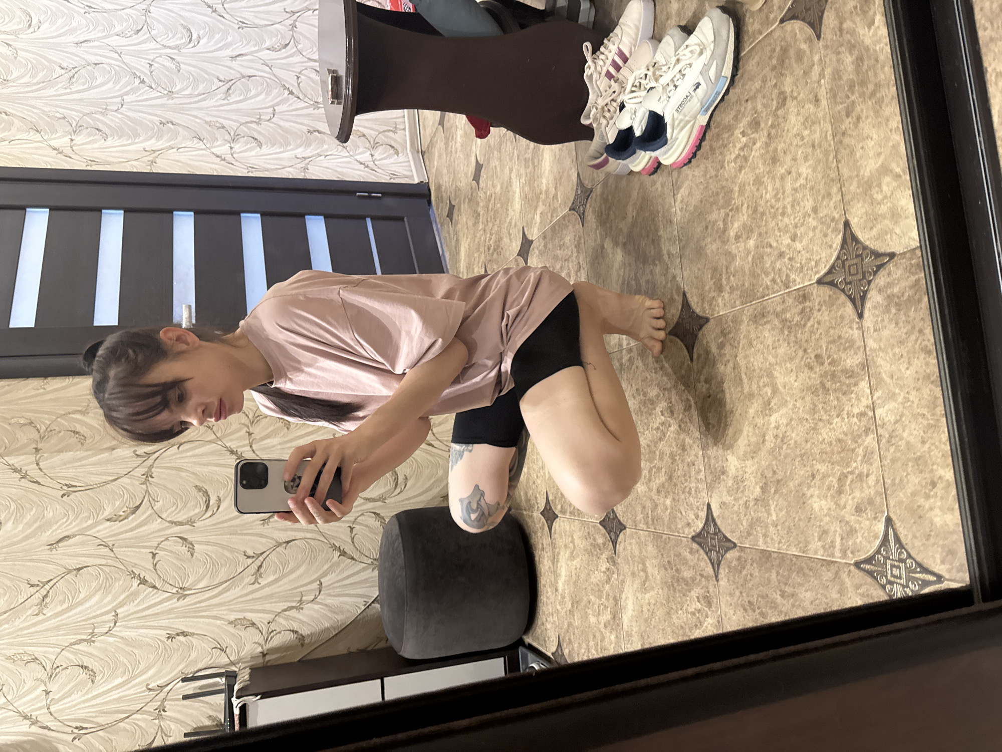A woman is crouching in what appears to be a bathroom, taking a photo of herself in a mirror. She's wearing a light pink t-shirt and black shorts. There's a black cushioned stool beneath her. The bathroom has patterned wallpaper and tiled flooring with star-shaped patterns. A pair of white sneakers are visible near a brown table leg. The image seems to be taken inside a private residence in an unknown city. There is no way to verify the location in Rostov-on-Don, Russia, or any other city based on the image alone.
