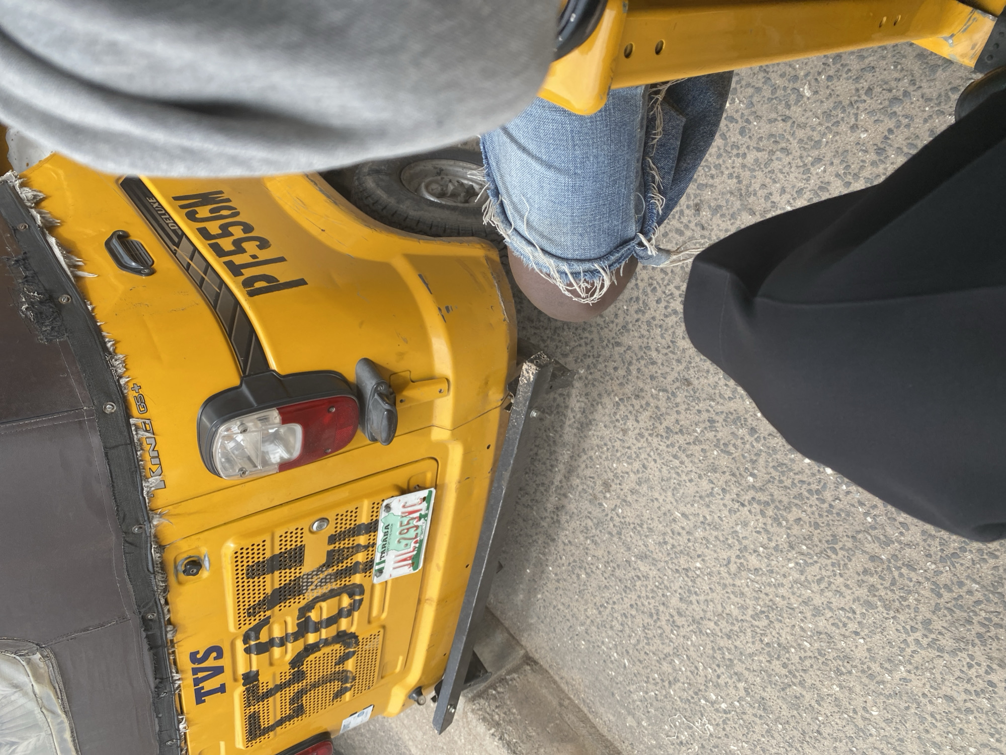 The image shows the rear of a yellow auto rickshaw with the license plate "JAL-295YC TARABA". The auto rickshaw has "TVS" and "PT55GM" written on it. The scene is outdoors on a paved road, possibly in Jalingo, Nigeria, given the license plate. The lower half of the image shows a person's legs wearing ripped denim jeans and a black garment, indicating they are a passenger in the auto rickshaw. The overall condition of the auto rickshaw appears worn.
