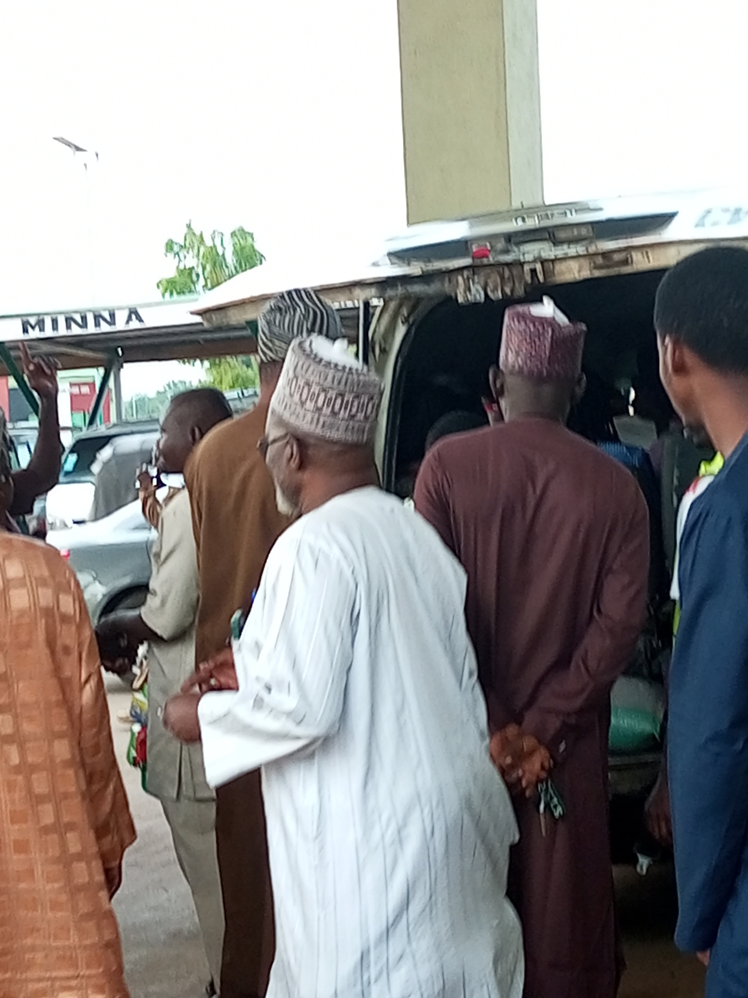 The image depicts a group of men gathered outdoors near a vehicle. The background shows a white structure with the word "MINNA" visible, suggesting a possible location or destination. One man in the foreground wears a white patterned garment and a matching cap. Another man to his right wears a maroon robe and a patterned cap. A third man, partially visible on the left, is in a tan garment. In the background, a silver car is parked. The overall atmosphere appears to be a public gathering.