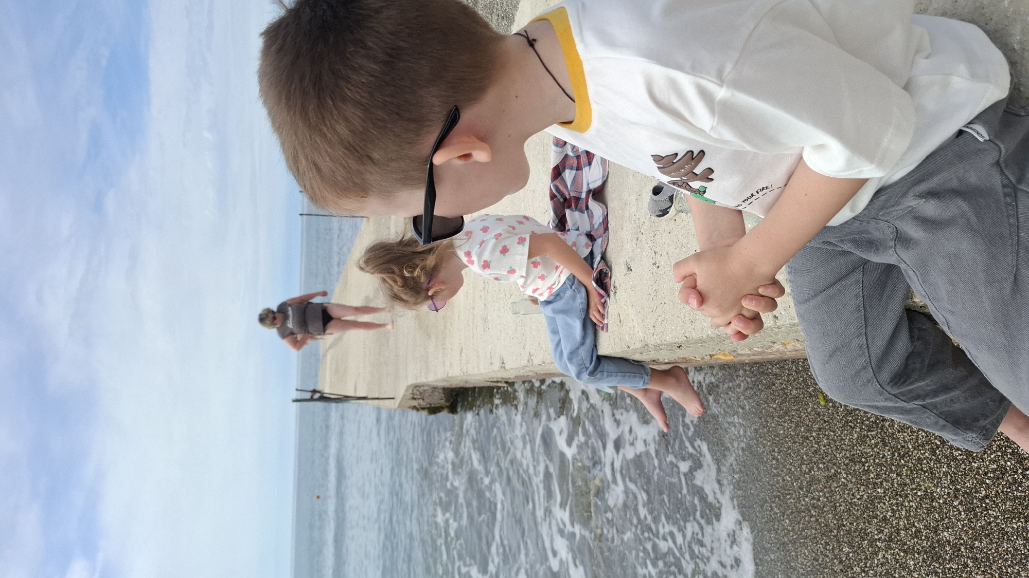 The image captures a serene coastal scene on a bright, likely overcast day, featuring a concrete pier extending into the sea, with a pebble beach alongside.
In the foreground on the right, a young boy with light brown hair is visible from the side, wearing a white t-shirt with a yellow collar and a graphic, below which partially reads "LIGHT UP YOUR FIRE!". He wears grey pants, and dark sunglasses are pushed up on his head. His hands are clasped together.
Next to him, a younger girl with light brown hair is crouching on the concrete pier, looking intently downwards towards the water. She is dressed in a white t-shirt with a floral pattern, light blue jeans, and small purple-tinted sunglasses. Her bare feet are dangling close to the water's surface, which shows gentle ripples and small waves. A plaid shirt is draped on the concrete barrier beside her.
Further down the pier on the left side of the image, an adult with short hair, wearing a grey t-shirt and dark shorts, stands casually. The expansive sea stretches out under a partly cloudy sky, with the overall atmosphere suggesting a relaxed, leisurely day by the water.