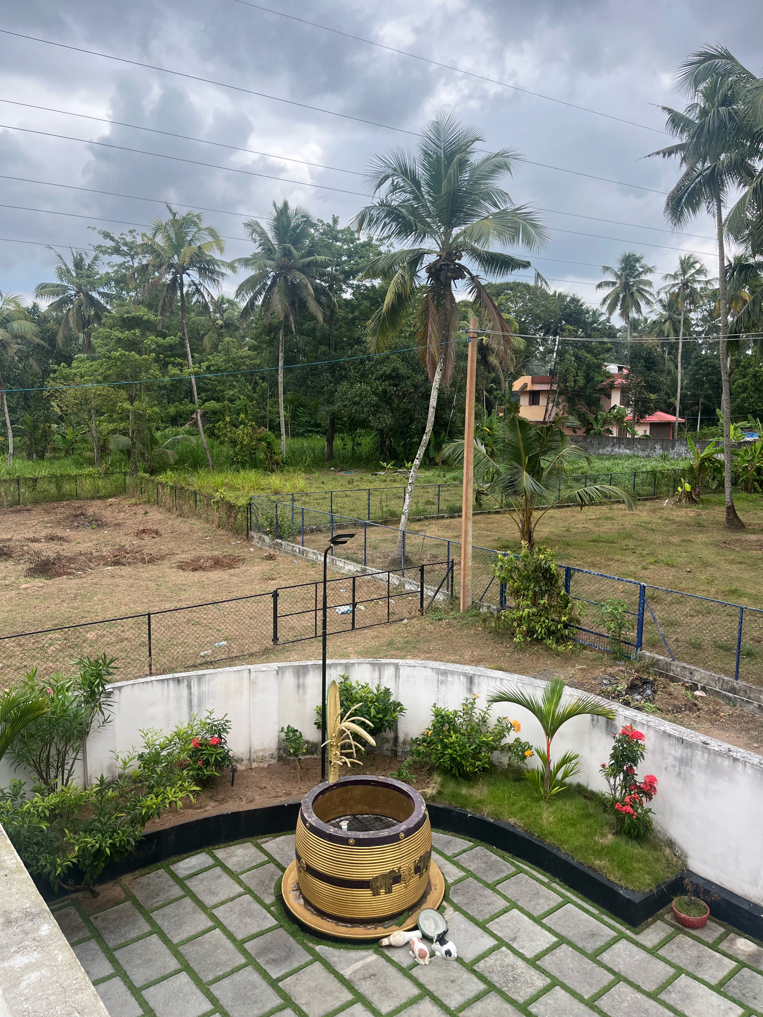 The image shows a view of a backyard in Kattanam, India, on a cloudy day. In the foreground, a paved patio area with square concrete tiles and artificial grass in between is visible. In the center of the patio is a large, decorative planter resembling a barrel, with gold and brown stripes and an embossed elephant design. Adjacent to it, a small garden bed contains various plants, including a small palm tree and flowering bushes with red and yellow blooms.
Beyond the patio, a white, low wall encloses the garden area. A chain-link fence with blue poles separates the yard from a larger, undeveloped grassy area. In the distance, more chain-link fencing encloses a section of dry, uncultivated land. Further back, a dense line of lush green trees, primarily palm trees, forms the background. Two houses with red tiled roofs are visible peeking through the trees. Overhead, power lines stretch across the cloudy sky. The scene appears to be a residential property with a mix of developed and undeveloped outdoor space. There are no people or animals visible in the image.