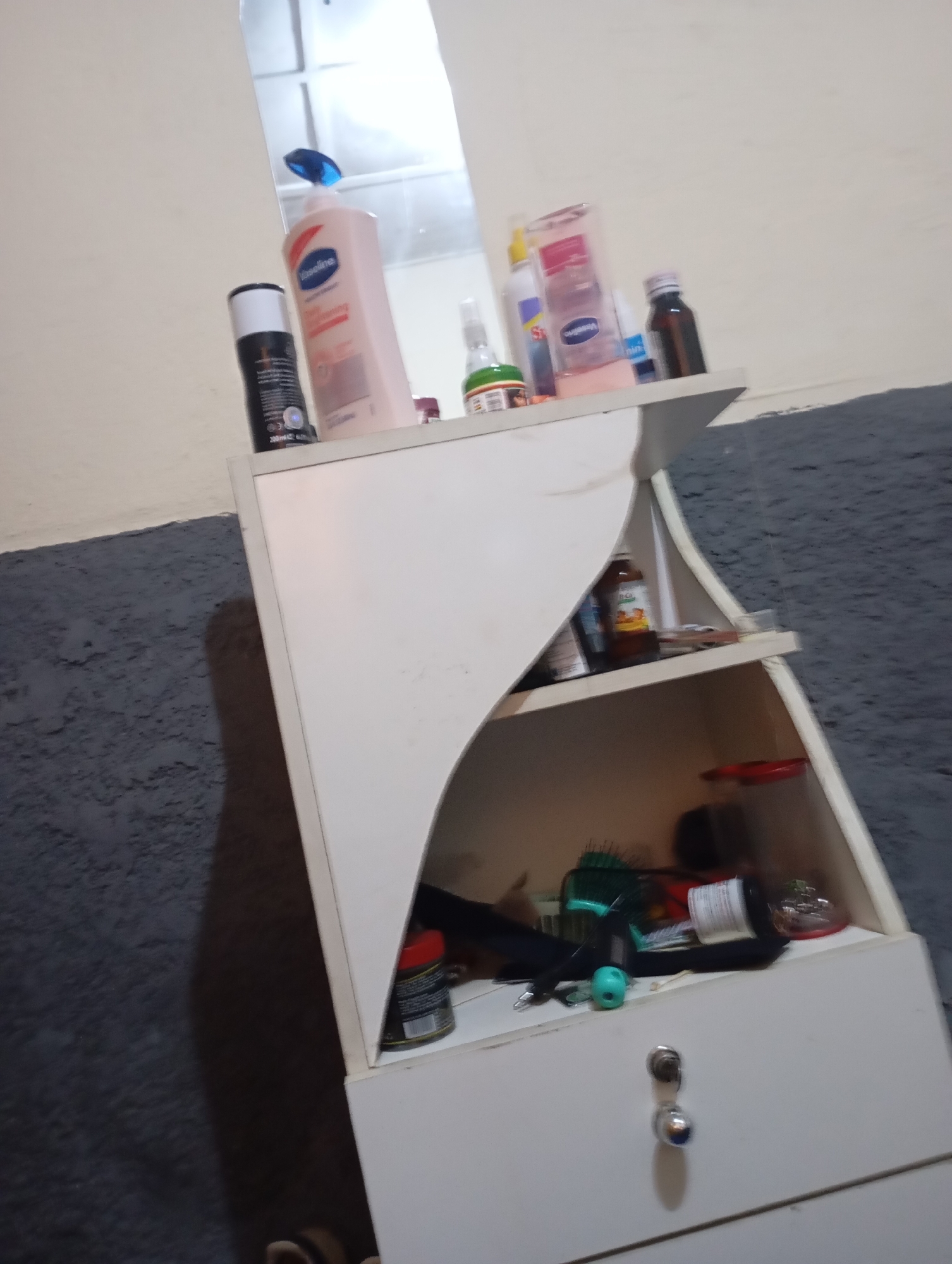A waist-high, white, two-tiered shelf unit with a curved cutout on the left side is positioned against a textured, dark gray wall. The top shelf holds several toiletries, including a can of black hairspray with a white cap, a large bottle of pink Vaseline lotion, a small clear bottle with a green cap, a rectangular pink bottle, and a small brown bottle. The lower shelf contains various items, such as brushes, bottles, and a red object. Below the shelves, a white drawer with two silver knobs is visible. The scene is lit by an overhead light, casting shadows. The background is a light-colored wall and a reflection in a mirror above the shelf unit.