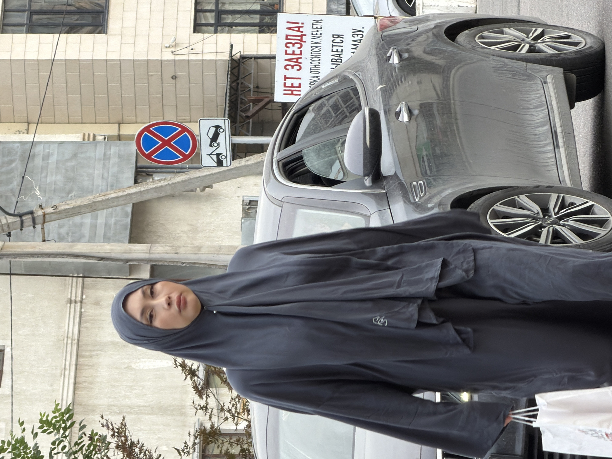 A woman wearing a dark gray hijab and a matching modest outfit walks past a parked dark gray sedan. She is carrying two white shopping bags in her left hand. The car has tinted windows and chrome door handles. In the background, a street sign with a red circle and a blue cross indicates no parking, and below it, a sign depicts a tow truck towing a car. Another larger white sign with red text that reads "НЕТ ЗАЕЗДА!" (No Entry!) is visible further to the right, with additional text below it. The scene appears to be on a city street with buildings and a utility pole in the background.