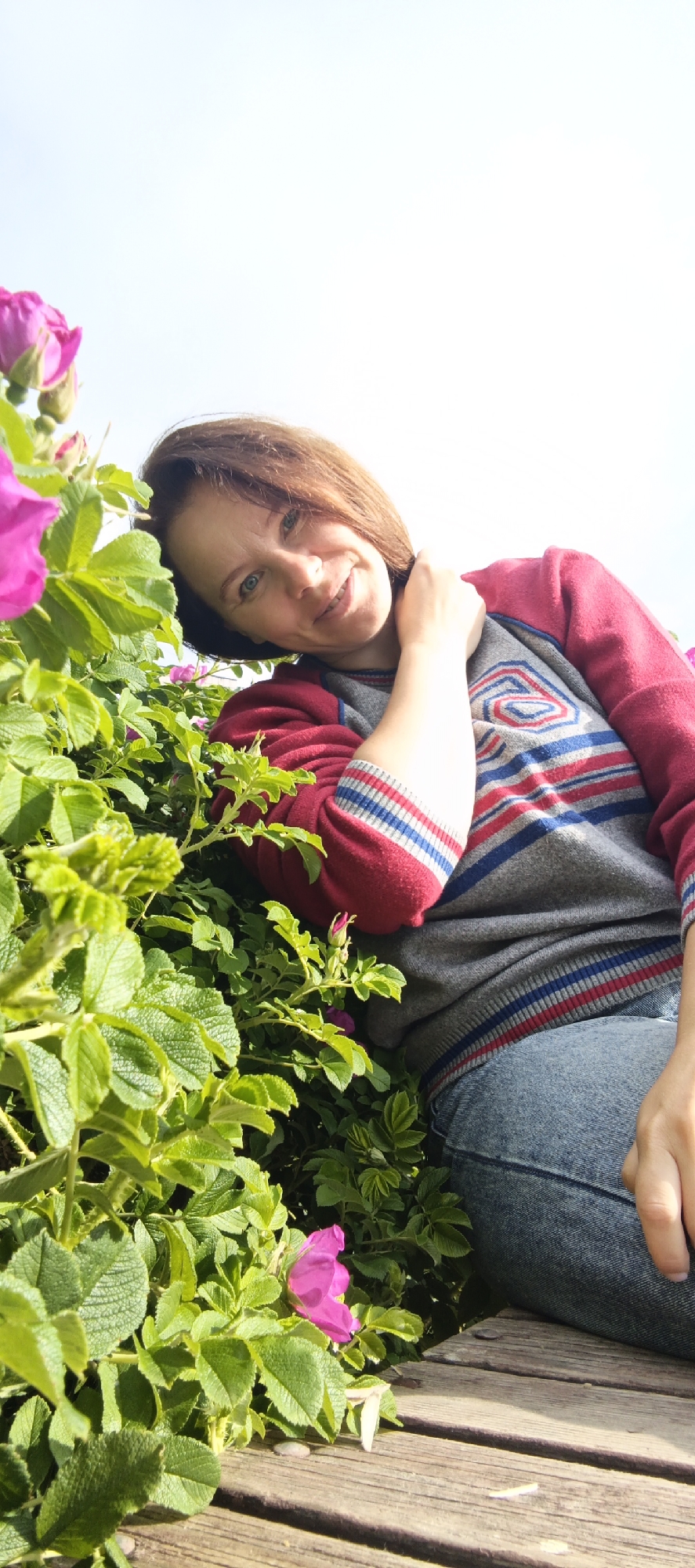 A woman with shoulder-length brown hair and blue eyes smiles at the camera, leaning her head on her hand and touching her neck. She is wearing a red and gray striped sweater and blue jeans. She is positioned among lush green foliage and pink flowers, with a wooden surface visible in the foreground. The background is a bright, overcast sky. The image appears to be taken outdoors during the day. The specific location in Chaikovsky, Russia is not identifiable from the visual information provided.