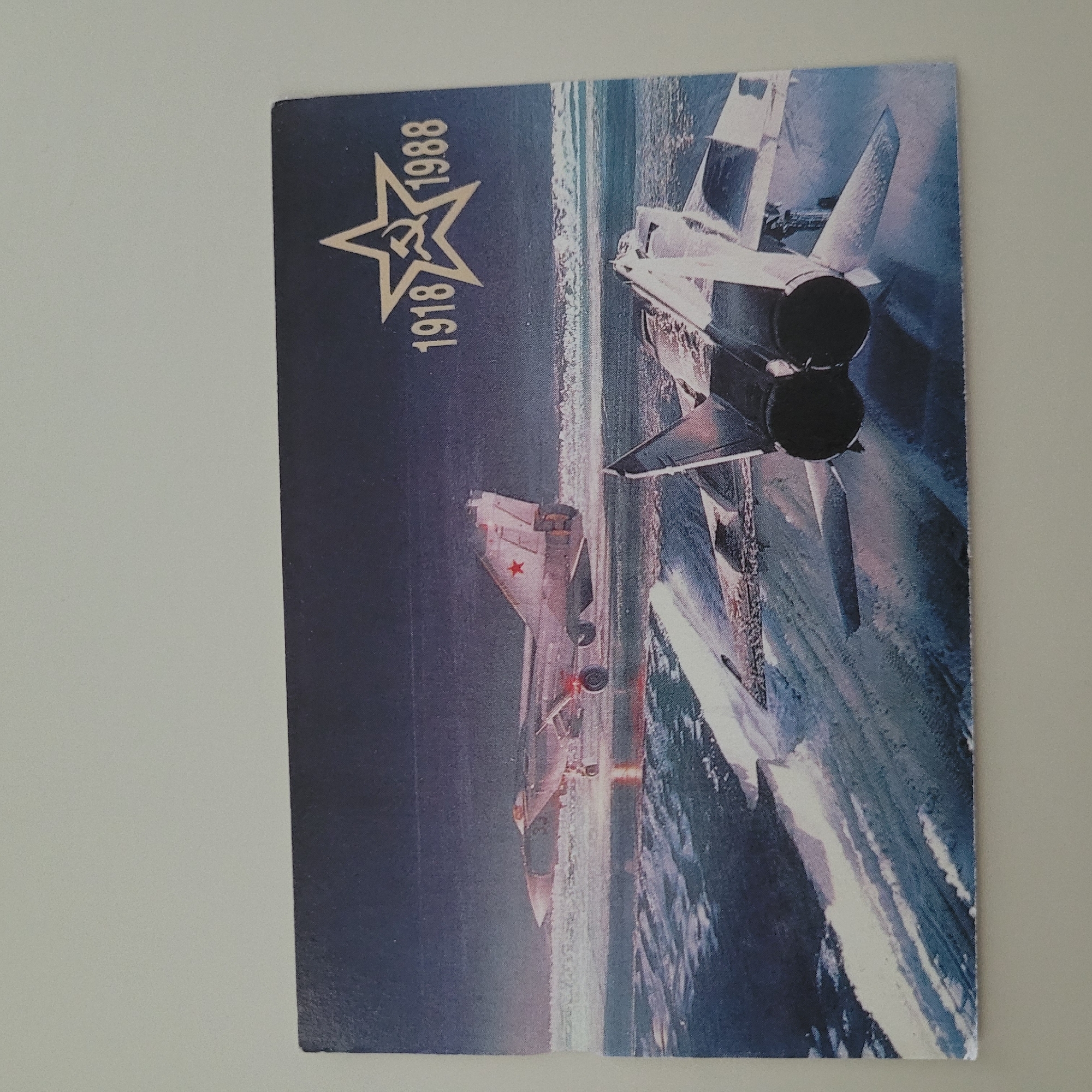 The image is a postcard depicting two Soviet-era fighter jets. One jet, a MiG-29, is in the foreground, viewed from behind, showing its twin engines and delta wings. Another jet, a MiG-21 with a red star insignia, is in the background, appearing to be on a runway or water surface. A stylized gold star with a hammer and sickle inside is visible in the upper right corner, along with the numbers "1918" and "1988." The setting appears to be a twilight or nighttime scene with water and reflections. The postcard is not located in Jūrmala, Latvia; it is a depiction of military aircraft.