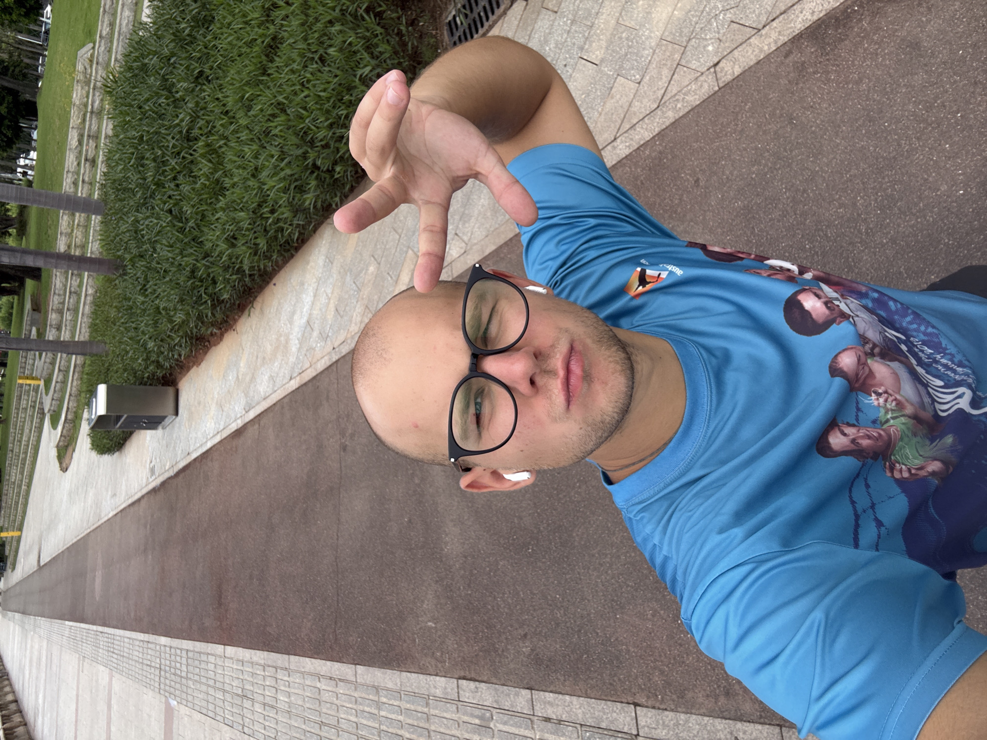 A man with a bald head, wearing glasses and a blue t-shirt, is taking a selfie outdoors. He has earbuds in both ears and is making a peace sign with his right hand. The t-shirt has a graphic design with images of people and text. The background shows a paved walkway, a grassy area with bushes, and some trees. There is also a trash can visible. The lighting suggests it is daytime. The location is the city of Tianhe District, China.