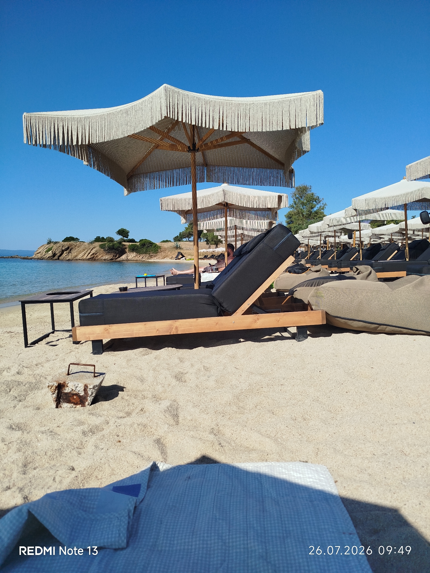 The image captures a sunny day on a sandy beach in Sithonia Municipal Unit, Greece. The scene is dominated by rows of lounge chairs and large, fringed umbrellas providing shade. In the foreground, a partially visible checkered blue fabric lies on the sand, casting a shadow. To the left, a dark-colored lounge chair with a wooden frame is prominent, accompanied by a small black table. Further back, a person is visible relaxing on a lounge chair under one of the umbrellas. The calm sea with clear blue water and a rocky coastline with greenery forms the backdrop, under a clear blue sky. The timestamp "26.07.2026 09:49" and "REDMI Note 13" are visible in the bottom left and right corners of the image.