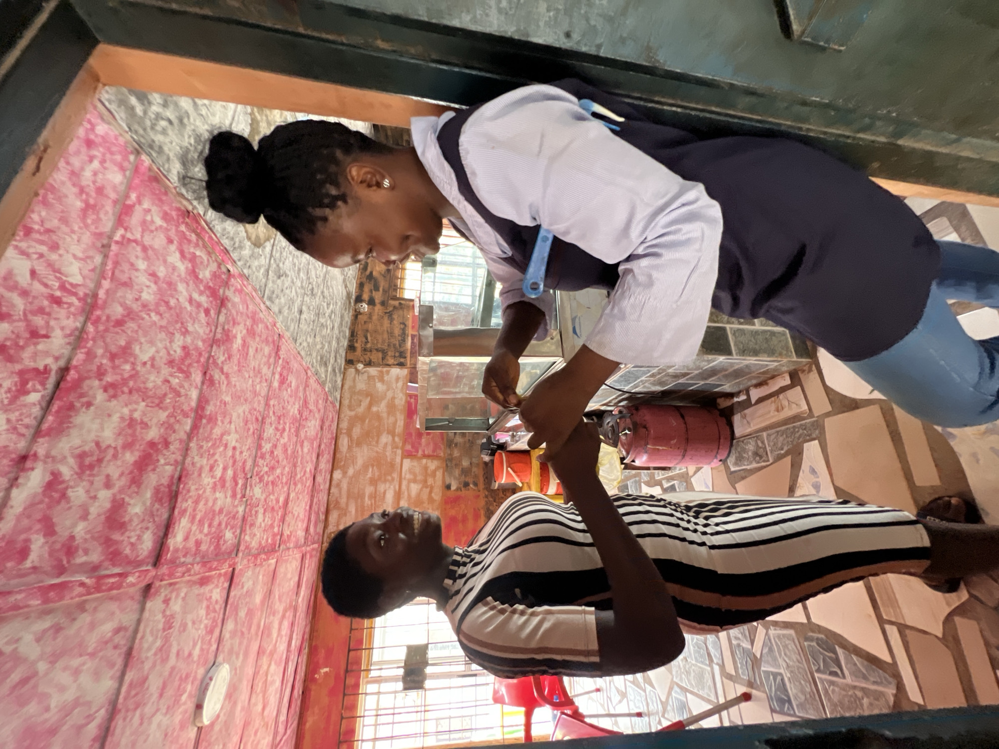 In Obosi, Nigeria, the image depicts two women interacting within an interior space. One woman, wearing a striped dress, stands while the other woman, in a light blue, collared shirt with a dark vest, is holding the first woman's hands. The background includes a textured pink and white wall, a tiled floor, and a gas canister. Additional objects include red chairs, visible through a window, and various household items. The environment appears to be a modestly furnished interior.