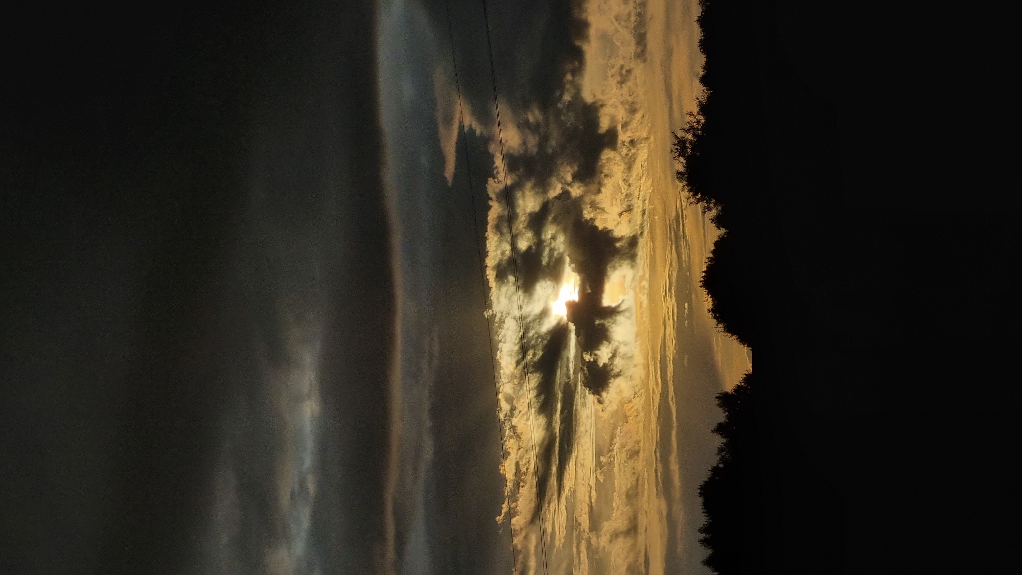 The image depicts a dramatic sunset over a silhouetted treeline in Vysokoe, Russia. The sky is filled with dark, heavy clouds, creating a sense of impending storm. However, shafts of golden sunlight break through the clouds, illuminating them from behind and casting a warm glow on the scene. Two thin, dark wires are visible stretching across the sky, suggesting their proximity to a power line. The foreground is dominated by the dark silhouette of dense trees, which form a stark contrast against the vibrant sky. The overall atmosphere is moody and intense, with a blend of darkness and light that is characteristic of a dramatic sunset.