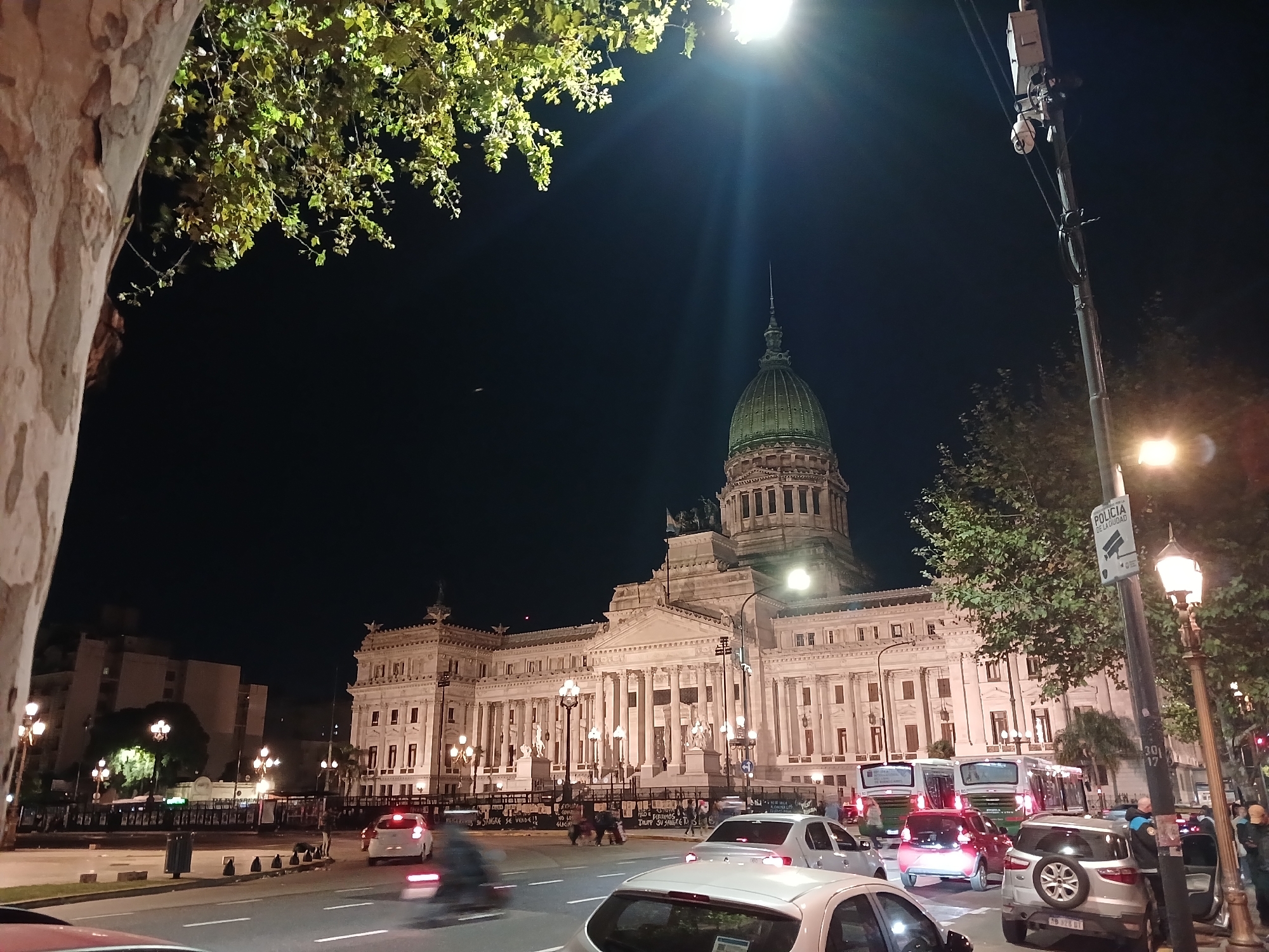 At night, a view of the Palace of the Argentine National Congress, also known as the Argentine Congressional Palace, is captured. The dome of the building is illuminated, accentuating its architectural details. In front of the Palace, a street scene is unfolding with moving traffic. A mix of cars and buses can be seen navigating the road. Pedestrians are also present, adding to the urban activity. A street sign with "POLICIA DE LA CIUDAD" is visible. The trees and streetlights lining the road further contribute to the vibrant urban environment.