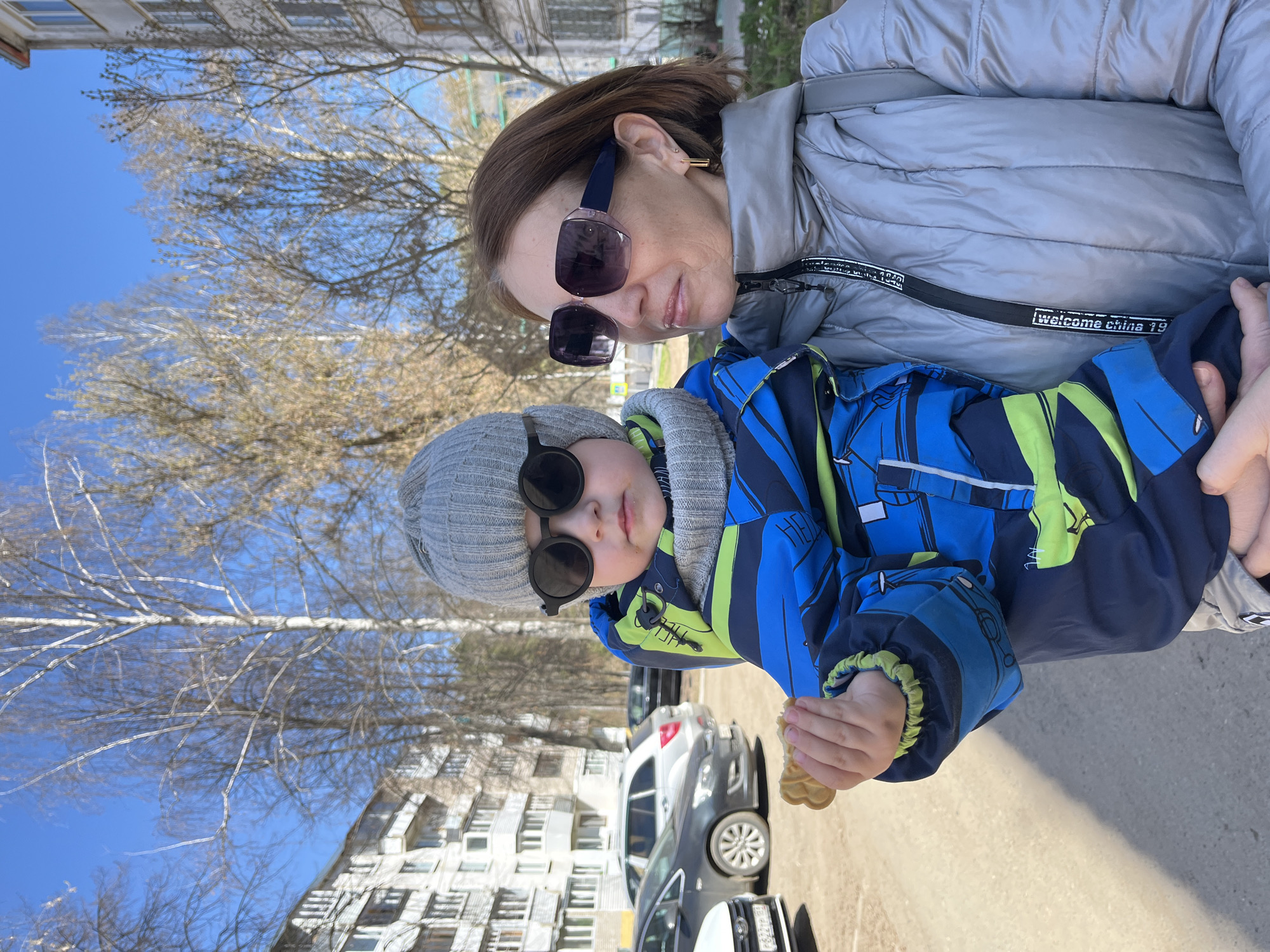 Here is a detailed description of the image:
**General Description:**
The image depicts a woman holding a young child outdoors. Both are wearing sunglasses. A building and cars are in the background.
**People:**
* **Woman:** Appears to be middle-aged, wearing a light gray puffer jacket and sunglasses.
* **Child:** The child is wearing a gray beanie, sunglasses, and a blue and green winter jacket. They are holding something small in their hand.
**Environment:**
* **Setting:** A paved outdoor area, likely a sidewalk or parking lot.
* **Building:** There is a multi-story building in the background with many windows.
* **Weather:** Seems to be a sunny day.
* **Cars:** Several cars are parked in the background.
**Objects:**
* Sunglasses (on both the woman and child)
* Winter clothing (jacket, hat)
* Cars
* Building
**Location:**
There is not enough information to confirm the exact location in the city of Chaikovsky, Russia.