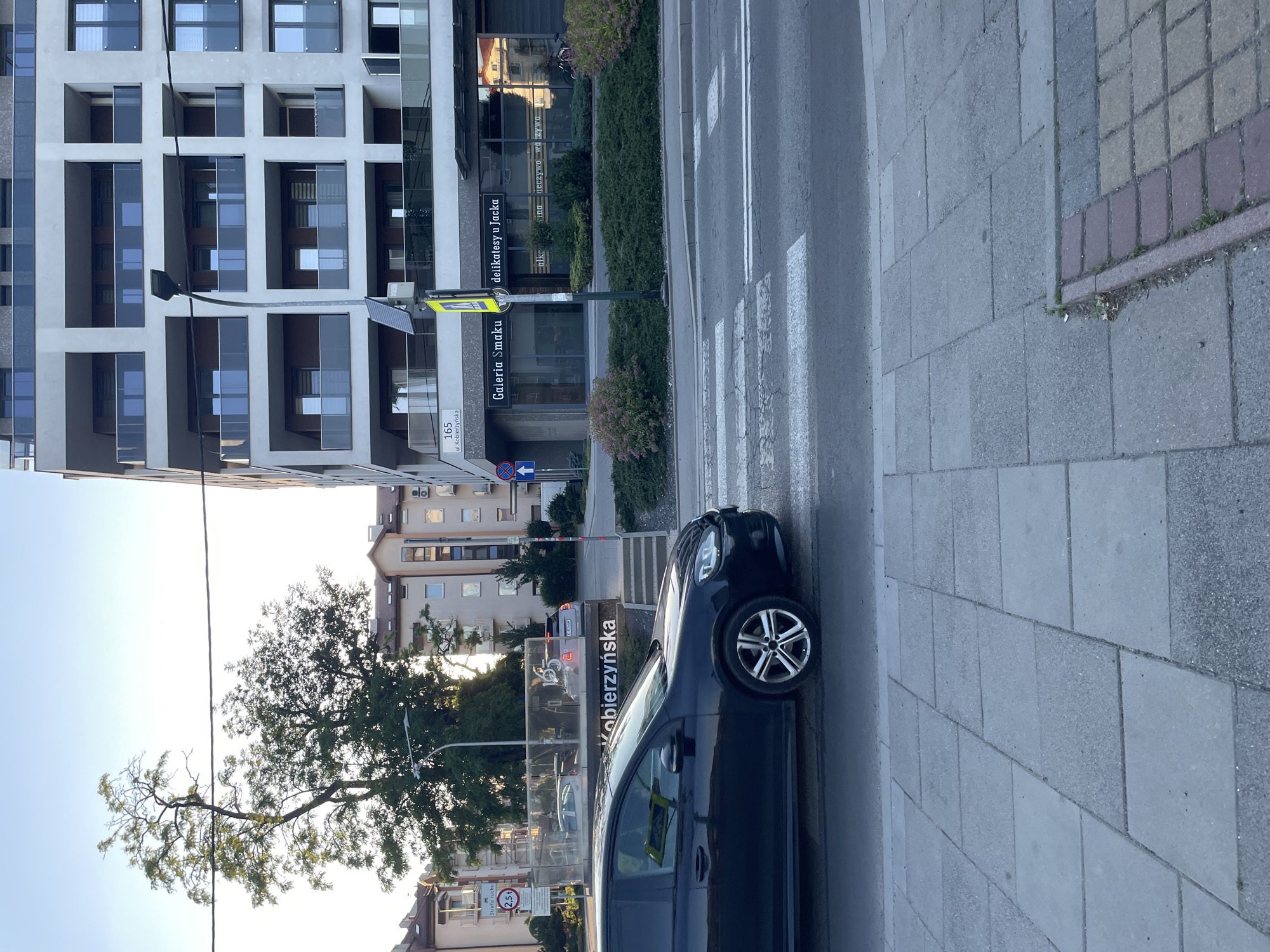 The image depicts an urban street scene on ul. Kobierzyńska, near number 165, in Kraków, Poland, during the late afternoon or early evening. In the immediate foreground, a grey tiled sidewalk is visible, bordered by a curb and a section of reddish-brown paving stones on the bottom right. A white-striped pedestrian crossing extends across the asphalt road from left to right.
A dark-colored, four-door Volkswagen Golf hatchback is positioned on the left side, partially on the pedestrian crossing, facing right. Its front wheels are on the crossing, and the driver's side is visible. In the mid-ground, a dark green street light pole stands at the center, featuring a yellow triangular pedestrian crossing sign and a small solar panel mounted below the lamp.
To the right of the street light, a modern multi-story apartment building with a light-colored facade and dark-framed windows with glass balconies is prominent. The ground floor of this building houses commercial establishments, indicated by signs reading "Galeria Smaku" (Gallery of Taste) and "delikatesy u Jacka" (Jacek's Delicatessen). A black street number "165" and "ul. Kobierzyńska" are visible on the building's facade above these shops. A small green median strip with bushes separates the road from the building. A bicycle is parked near the entrance of "delikatesy u Jacka."
To the left, behind the Volkswagen Golf, a glass-paneled structure, possibly a bus stop or public information display, has "Kobierzyńska" text across it. Through this structure, an SUV and another car are partially visible. Further in the background, several trees with green foliage and multiple older residential buildings with traditional pitched roofs and various windows are visible under a clear, bright sky. Overhead electrical wires stretch across the upper portion of the frame. Street signs, including a red circular sign with "2.5t" and "Strefa ruchu" (Traffic Zone), and a blue square sign with a white arrow pointing upwards, are also present on the left side of the street.