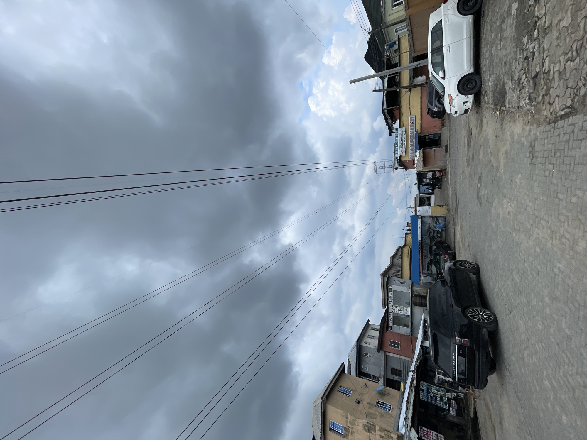 The image shows a street in Ado, Nigeria. The sky is overcast with heavy, dark clouds. Power lines stretch across the sky. Buildings line both sides of the street, with storefronts at street level. A white car and a black Range Rover are parked on the right side of the road. People can be seen walking in the distance.