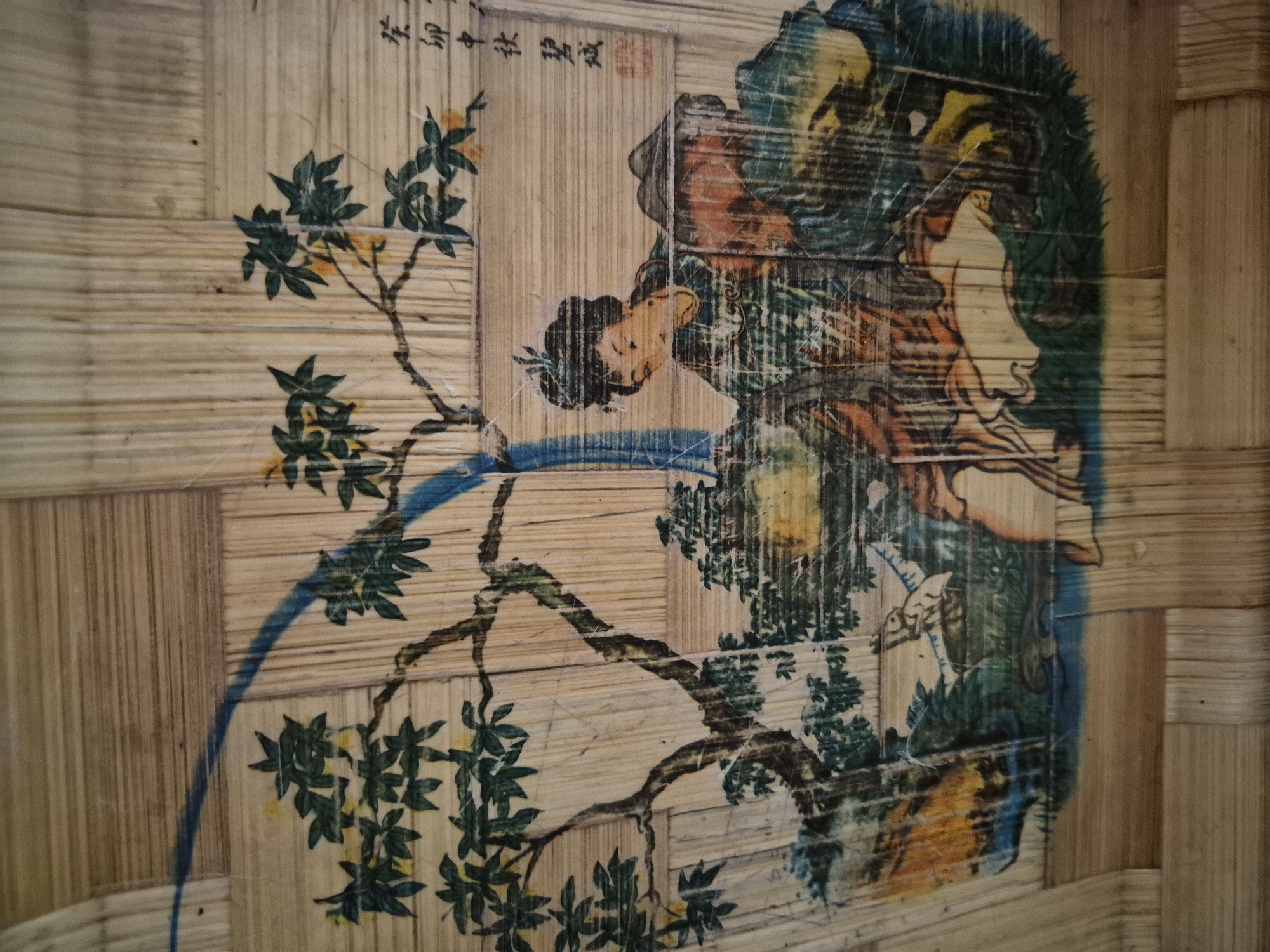 The image depicts a painted scene on a woven bamboo surface, possibly a fan or a decorative panel. The painting features a tree with green leaves and yellow fruits, a blue curved line suggesting a river or stream, and two figures. One figure, a woman with dark hair in an updo, wears a green garment. She is interacting with another figure, partially obscured, wearing a conical hat and an orange-red garment. The scene is rendered in a traditional East Asian style. The woven bamboo creates a textured background, with individual strips running vertically and horizontally. In the upper right corner, there are Chinese characters, possibly a signature or inscription, and a red seal. The painting appears to be aged, with visible wear and tear, and some scratching on the surface.
There is no information in the image that would allow for its location in the city of Vanadzor, Armenia.