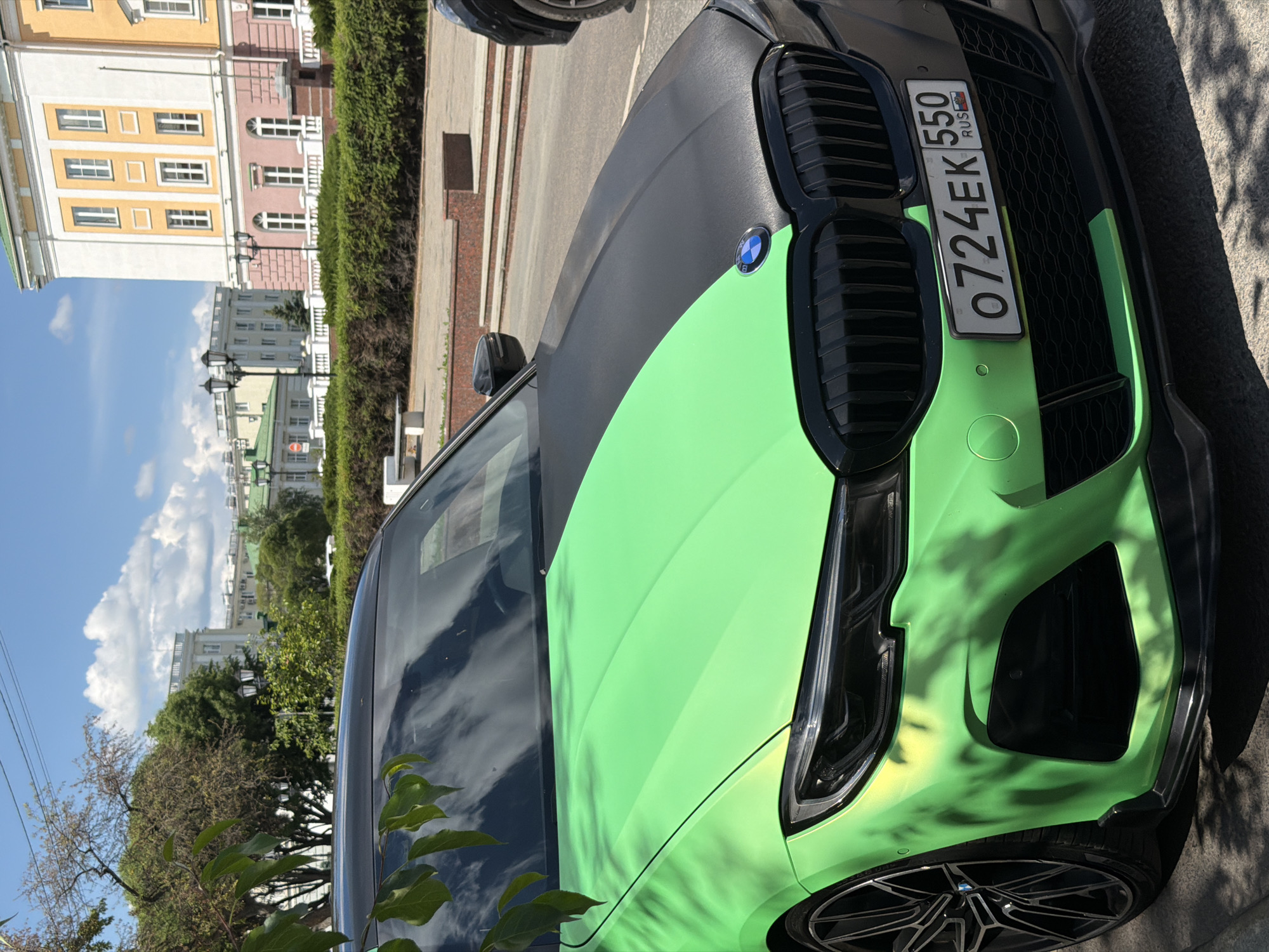 A close-up, eye-level, full-shot photograph depicts the front of a black and neon green BMW car parked on a paved surface in Izhevsk, Russia. The car's hood is split vertically between the two colors, with the driver's side predominantly neon green and the passenger's side matte black. The front grille and headlights are visible, as is the license plate, which reads "O 724 EK 550 RUS". In the background, a building with yellow and pink facades, white trim, and arched windows stands under a blue sky with scattered clouds. Green trees and bushes line the foreground and the area around the building. The scene is well-lit, suggesting it is daytime.