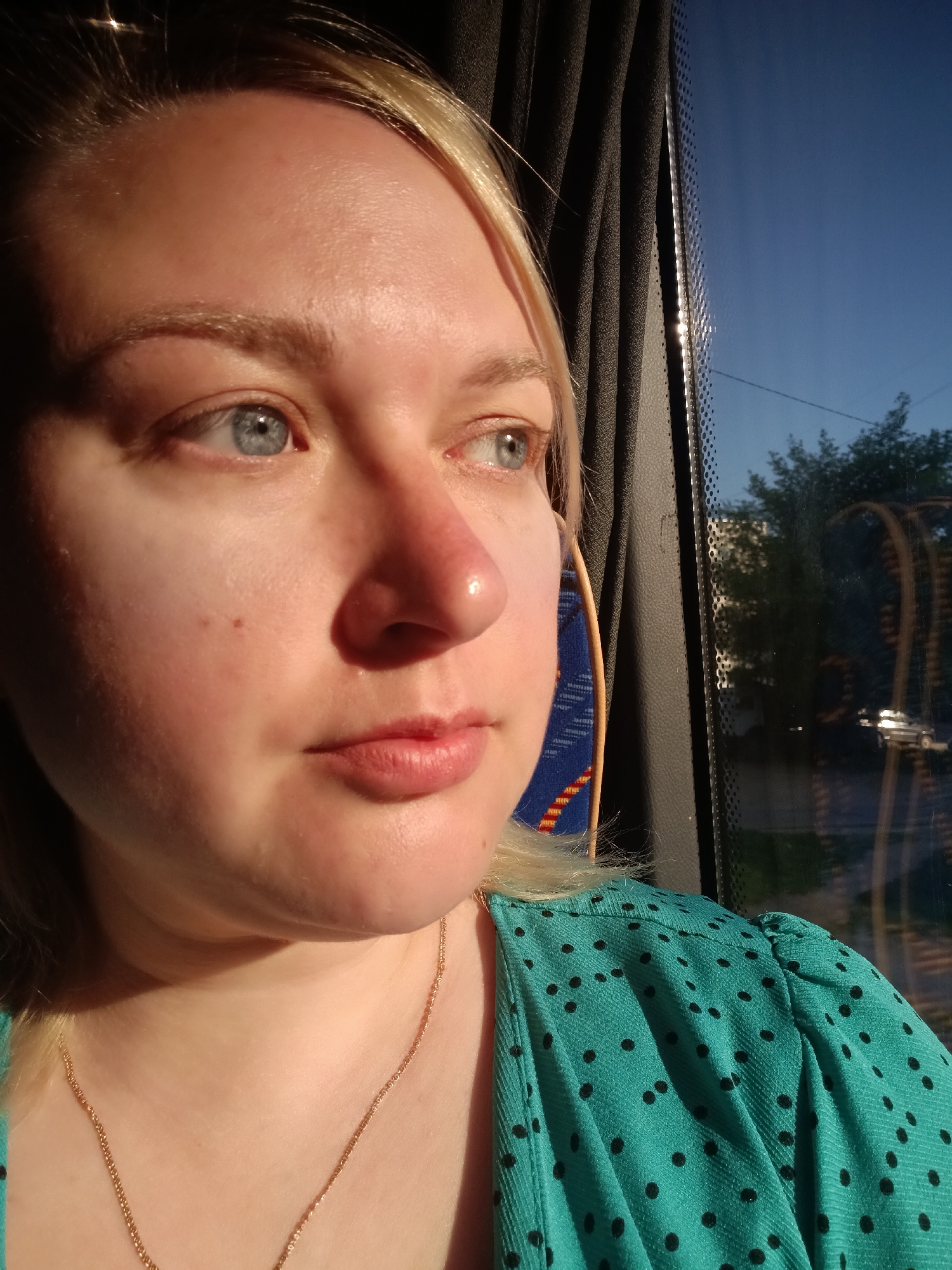 A fair-skinned woman with blond hair, blue eyes, and wearing a teal blouse with black polka dots and a gold necklace is looking out a window. The window is likely that of a bus or train, based on the interior paneling seen through the window. Outside the window, trees and a building can be seen. Considering the photo was taken in Chaikovsky, Russia, the buildings in the window are likely to be the typical Soviet-era apartment blocks. The sunlight suggests it is early morning or late afternoon. The woman's face is lit by the same sunlight, which causes a slight redness on her skin.