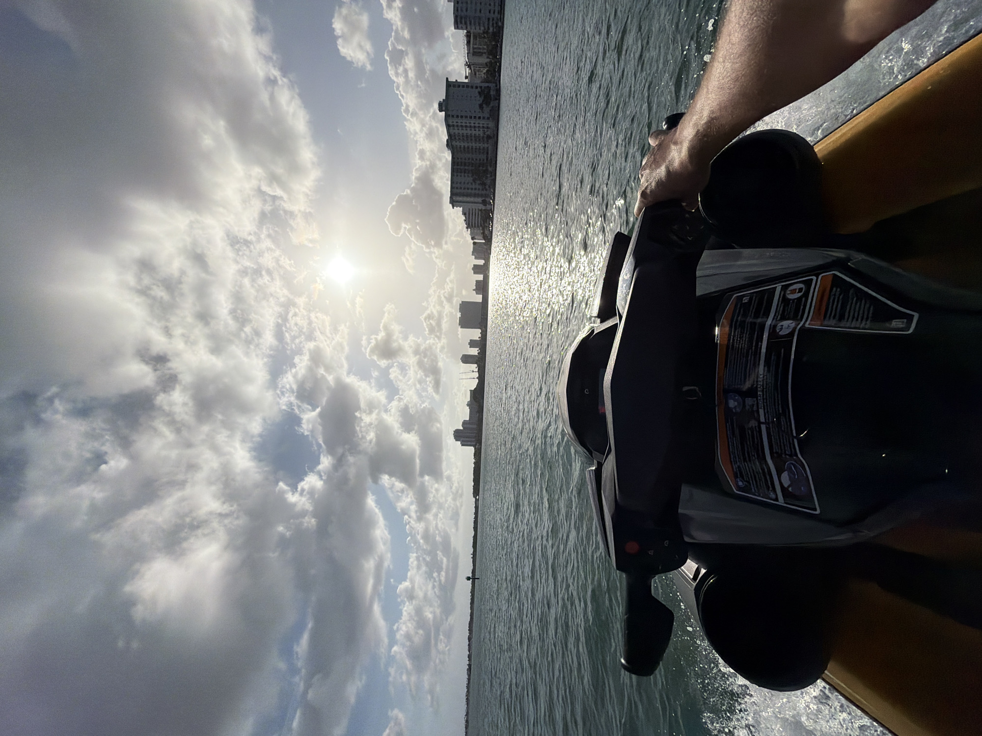 View from a personal watercraft (PWC) on Biscayne Bay, looking west towards the high-rise buildings of Bay Harbor Islands, Florida, USA. Sunlight pierces through clouds. A person's arm is visible in the upper right corner. The PWC's handlebars and dashboard are in the lower right foreground. Water is splashing around the PWC, indicating movement.