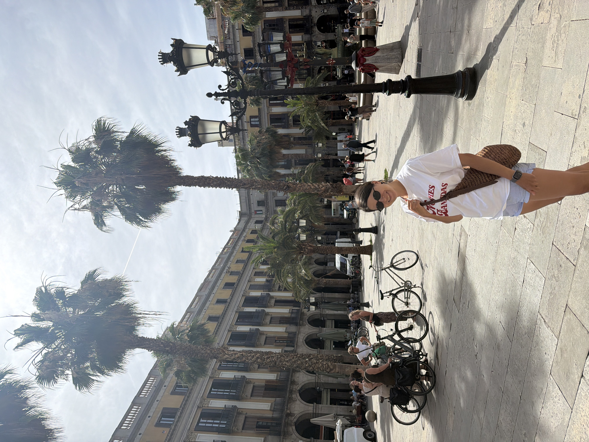 The image depicts a woman standing in a sunlit plaza in Barcelona, Spain, likely the Plaça de Catalunya, a major public square. She is positioned in the foreground, smiling at the camera, wearing a white t-shirt with red lettering, blue and white striped shorts, and sunglasses. A woven brown bag is slung over her shoulder. Behind her, several tall palm trees line the square, with ornate lampposts and a grand, multi-story yellow building with arched walkways visible in the background. Other people can be seen in the middle ground, some interacting with bicycles, while others stroll through the plaza. The paving of the square is made of large, rectangular stone slabs. The sky is partially cloudy.