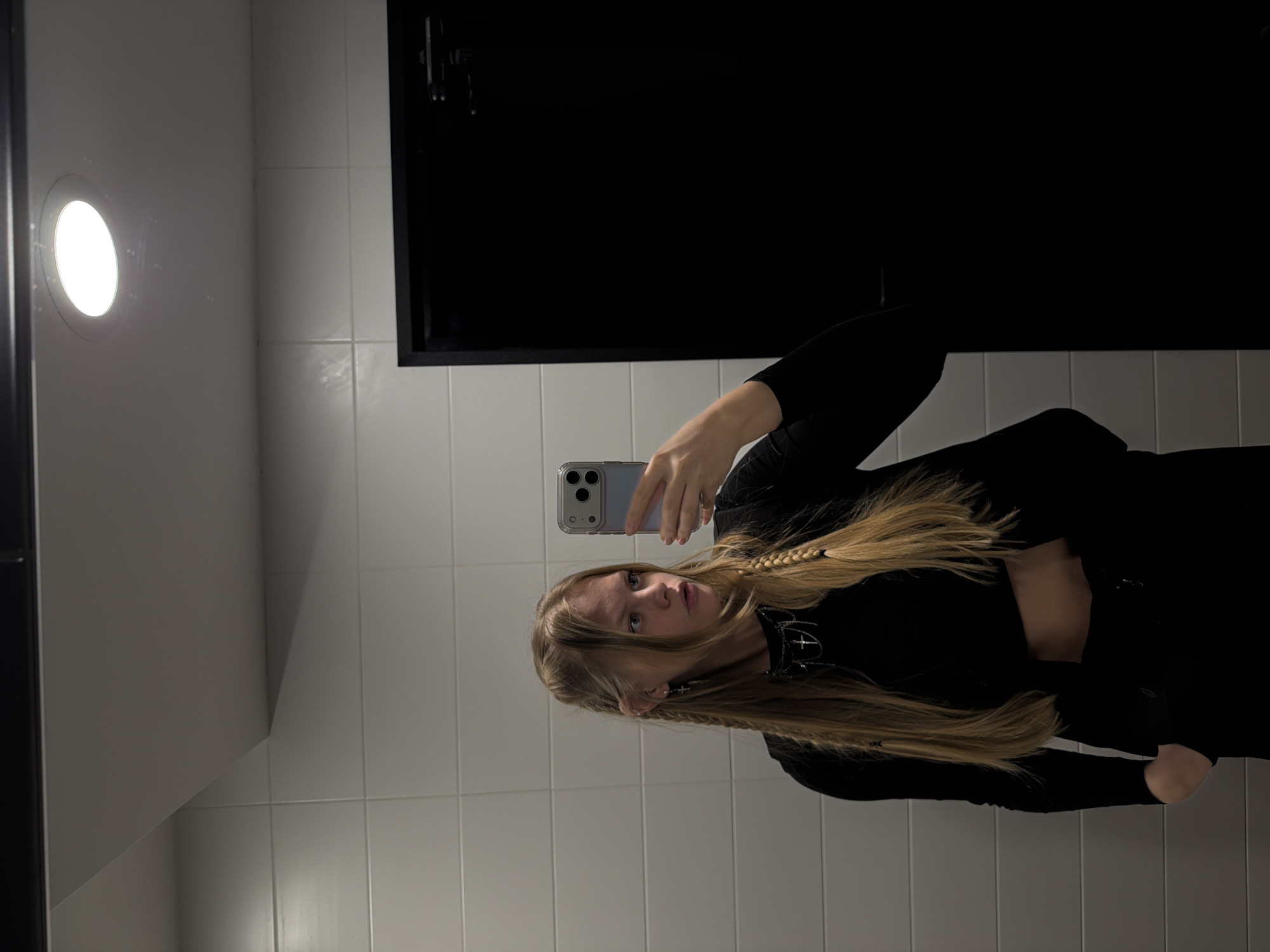 A person is standing in a bathroom taking a selfie. The walls are tiled and there is a light fixture in the upper left corner and a black rectangle, possibly a mirror or window, on the upper right. The person has long blond hair with a braid and is wearing a black outfit. The location cannot be confirmed as Izhevsk, Russia from the image alone.