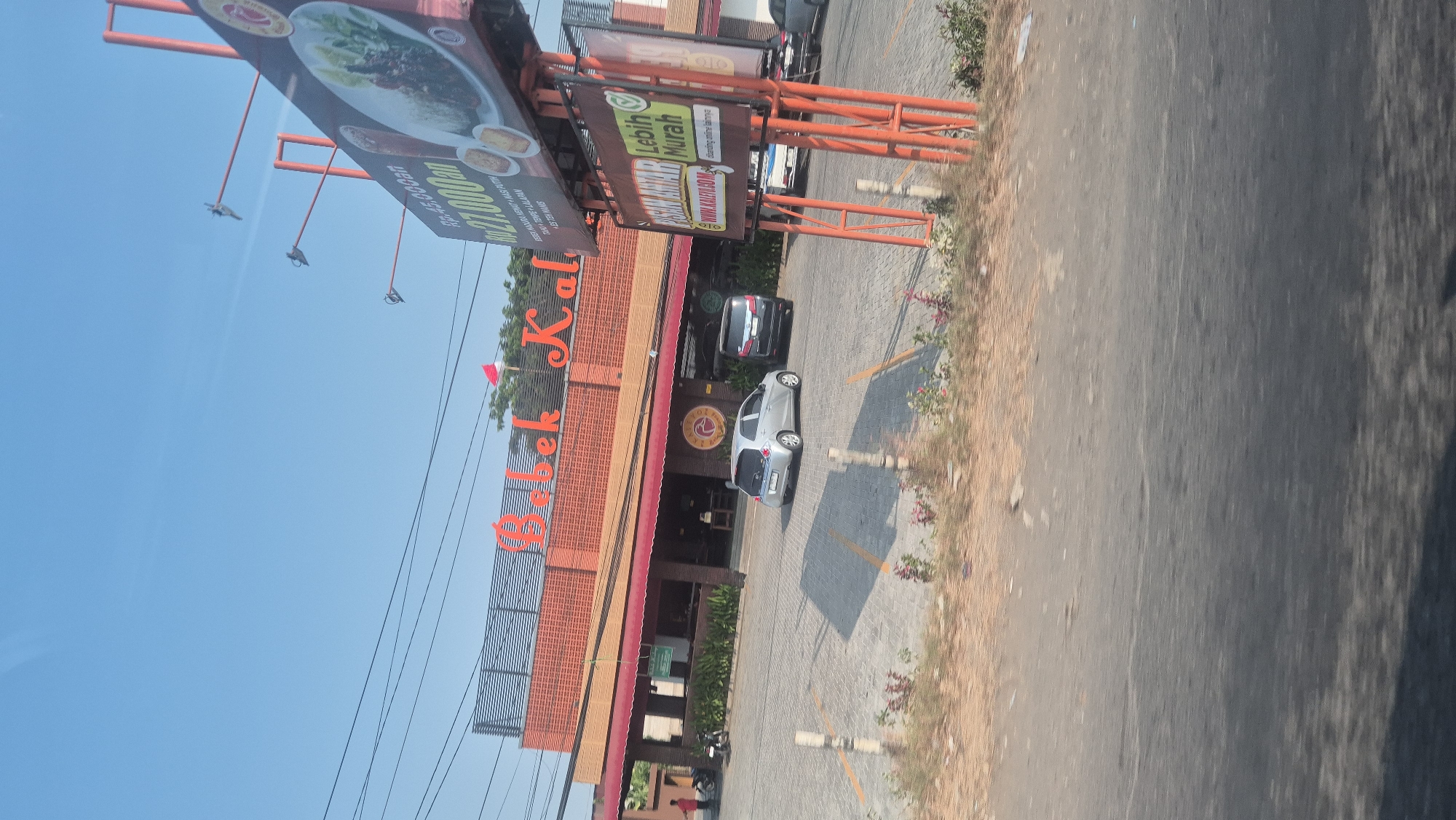 A low-angle, wide shot captures the exterior of a restaurant, likely "Bebek Kali", situated in Karawang, Indonesia, under a clear blue sky. The building features an orange brick facade with a red awning stretching across its front. Above the awning, large, orange, stylized lettering spells out "Bebek Kali". A small red and white Indonesian flag is visible on the rooftop. In front of the restaurant is a paved parking lot with several cars, including a silver hatchback and a black SUV. A tall billboard advertising food and prices stands to the right of the building, prominently displaying text and images of food items. The billboard has orange metal supports. A few people are seen near the entrance of the restaurant, and a motorcycle is parked on the left side. The foreground shows a road and a patch of dry, grassy ground with some small plants. Power lines are visible crisscrossing the sky.