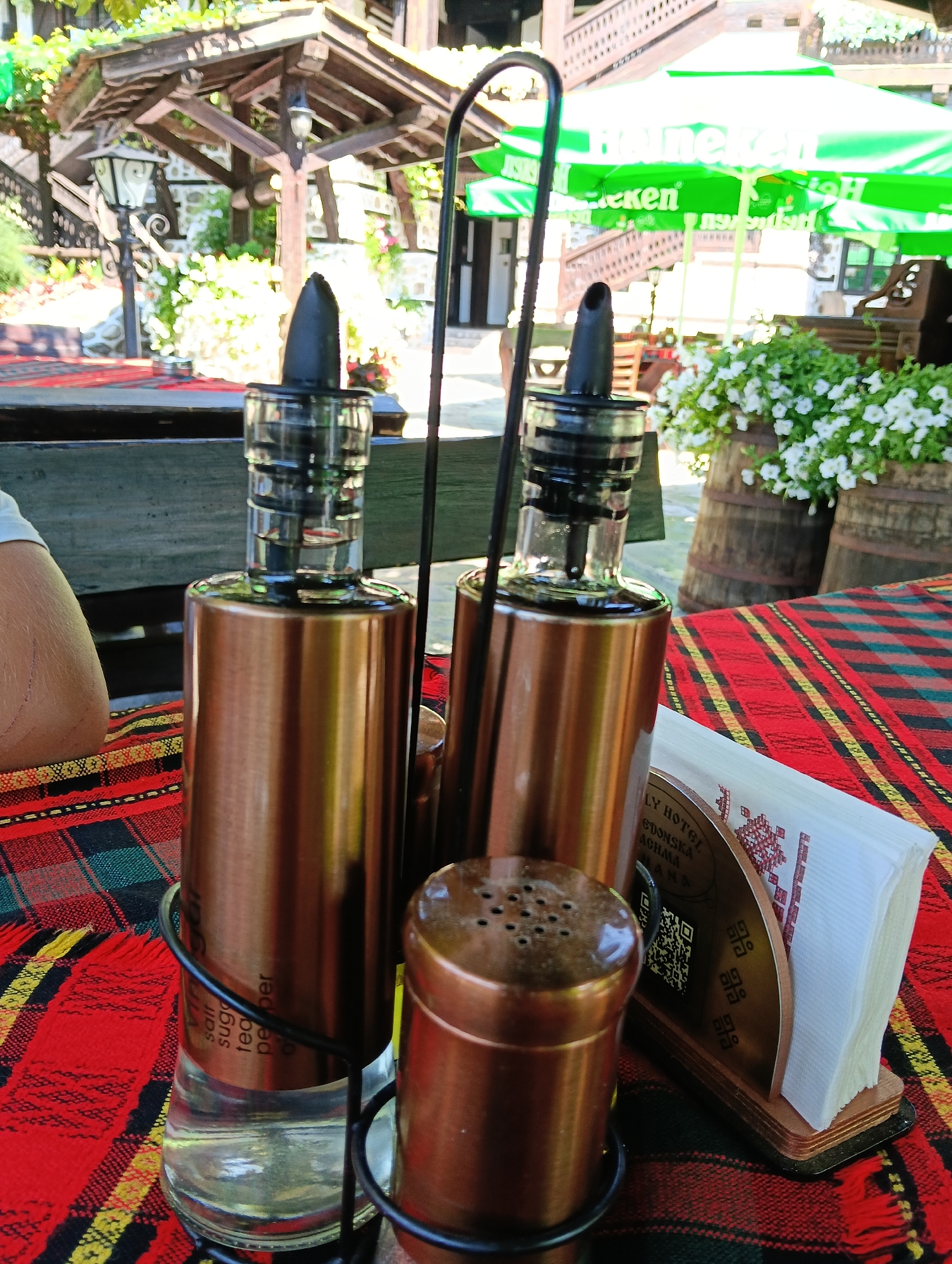 The image shows a close-up of a table setting at an outdoor establishment. A red, yellow, and green plaid tablecloth covers the table. On the table, there is a set of salt and pepper shakers and two oil or vinegar dispensers, all with copper-colored casings. To the right of the dispensers is a napkin holder with white napkins, featuring a decorative wooden base with what appears to be a QR code and text. In the background, there is a wooden gazebo with a thatched roof, a street lamp, and some greenery with white flowers. Two large green umbrellas with "Heineken" branding are visible in the upper right background. A wooden barrel with white flowers is also present. The overall scene suggests a restaurant or cafe environment. The presence of "LY HOTEL GDONSKA AGHALA," and "MANA" on the napkin holder and the traditional Bulgarian-style decor suggests this location is likely the restaurant "Aghalata" in Dobrinishte, Bulgaria.