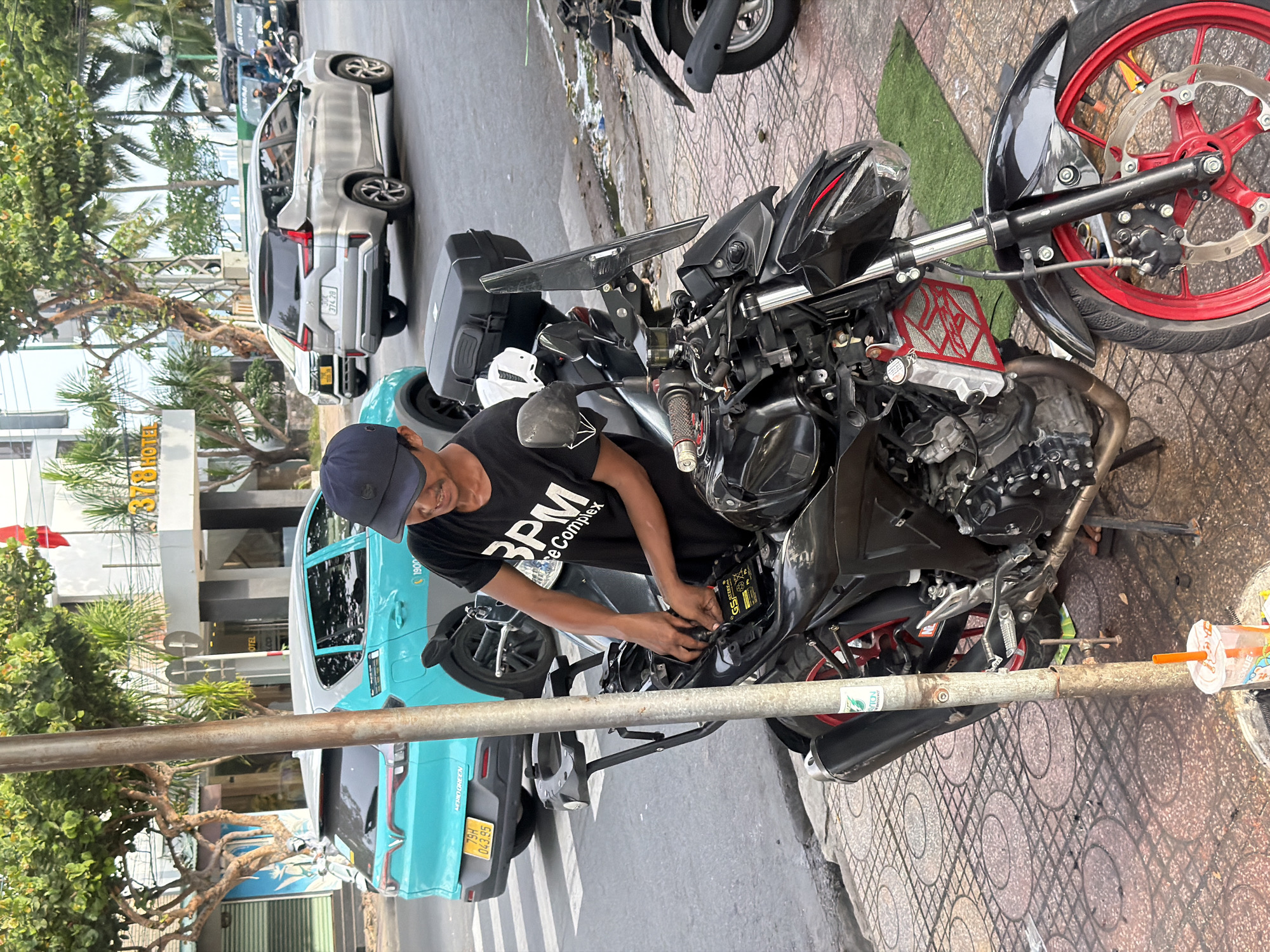 In Nha Trang Ward, Vietnam, a man wearing a black baseball cap and a black t-shirt with white lettering that reads "BPM Complete" is working on the battery of a black motorcycle. The motorcycle has red accents on its wheels and a black storage box mounted on the back. The man is focused on the task, with his hands inside the motorcycle's seat area. In the background, a bright teal car is parked on the street, and further down, a silver SUV is visible. The scene takes place on a paved sidewalk next to a street, with trees and buildings lining the background. A street pole stands prominently in the foreground, with a partial view of a trash can with a beverage cup and straw.