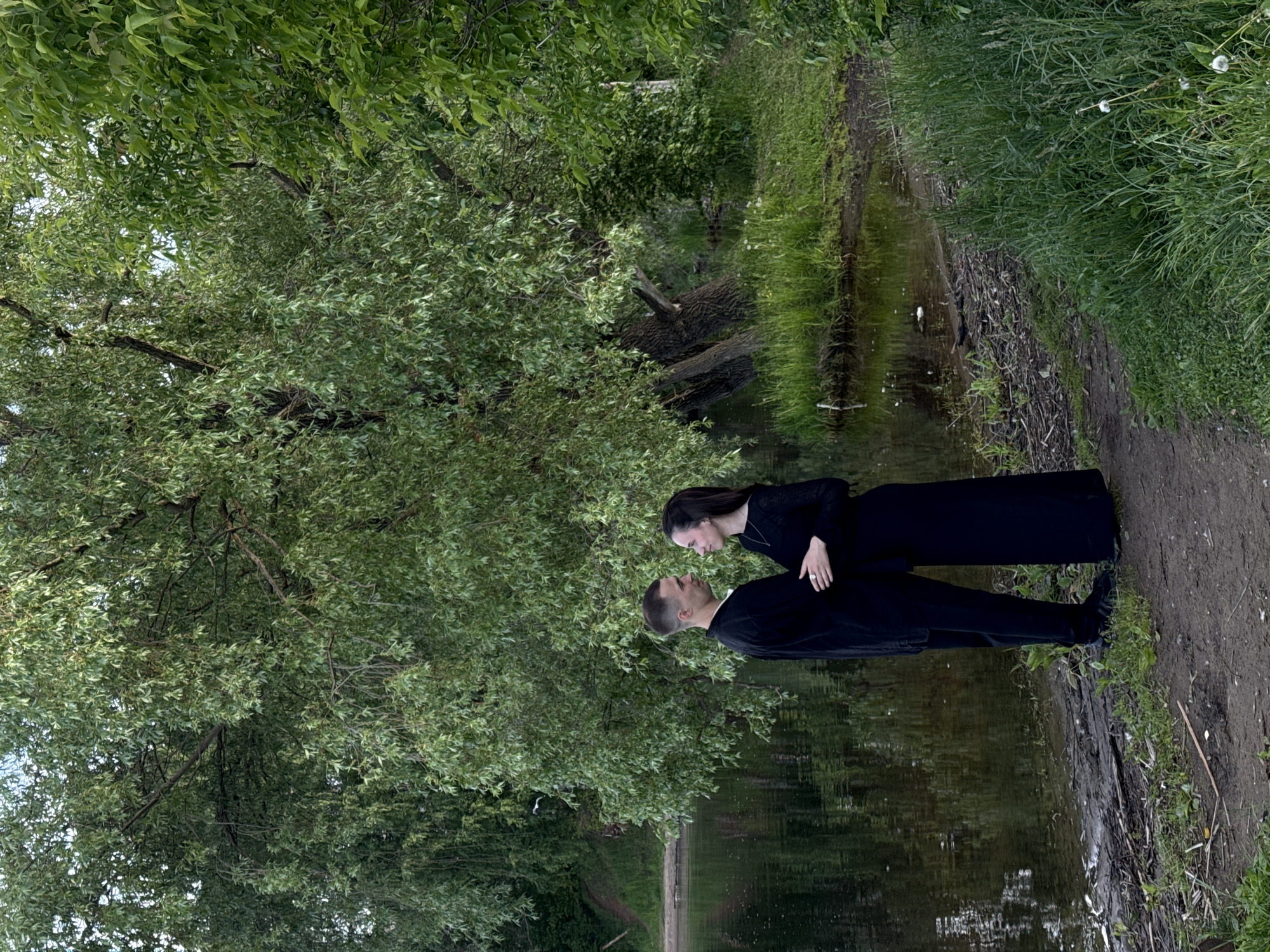 The image, when correctly oriented, captures a tender moment between a couple in a lush, green natural setting, likely in Chaikovsky, Russia. A man and a woman, both appearing to be young adults, stand facing each other near the edge of a body of water. They are dressed in dark, long-sleeved clothing, with the woman wearing a ring on her left hand. Her right hand is gently placed on the man's waist as they gaze into each other's eyes, suggesting affection and connection.
The scene is enveloped by dense, vibrant green foliage. Large trees with abundant leaves form a canopy overhead and extend along the left side of the frame, reflecting softly in the calm water. The body of water, possibly a pond or a slow-moving river, parallels a narrow dirt path that runs along the right side of the image. The path is bordered by tall, wild green grass.
The lighting is soft and diffused, indicating an overcast day or a deeply shaded area, creating a serene and tranquil atmosphere without harsh shadows. No specific man-made objects are prominently visible, and there is no discernible text within the image. The overall impression is one of a peaceful, natural environment providing a backdrop for the couple's intimate moment.