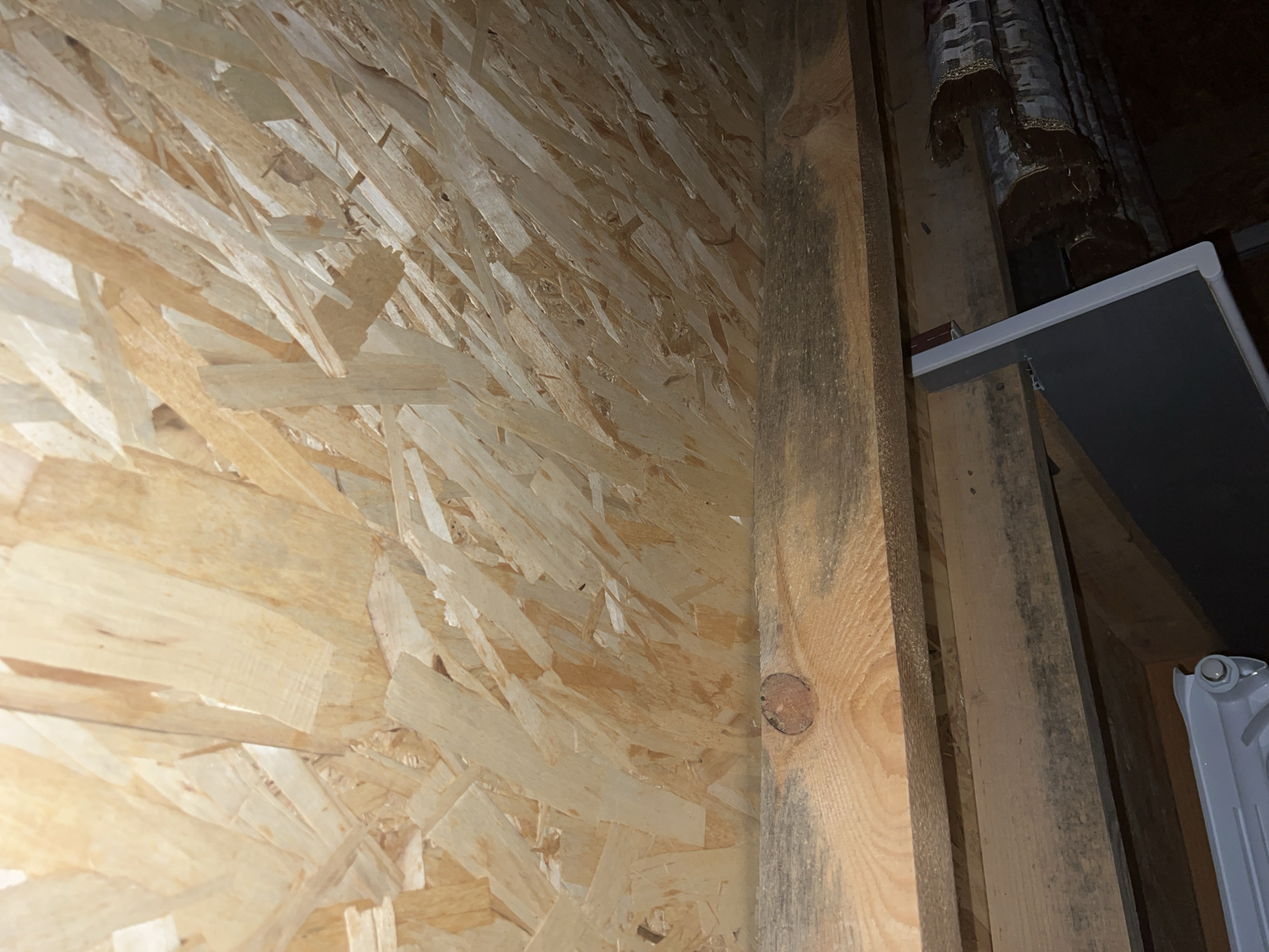 The image depicts an interior corner of a structure, likely under construction or in a state of disrepair, with a focus on the textured OSB (Oriented Strand Board) walls and wooden framing. The OSB is a light brown, layered material composed of compressed wood chips, with visible variations in color and grain. A substantial wooden beam, dark brown and showing signs of age with some discoloration and a visible knot, runs vertically along the corner. To the right of this beam, another, thinner wooden beam is present, also showing darker, possibly moldy, patches. Above and to the right, a portion of a white shelf is visible, supported by wooden bracing. Further in the background, a grey radiator-like object with a white knob is partially obscured, and above it, a patterned fabric, possibly a curtain or tapestry with fringe, hangs down. The lighting appears dim, suggesting an indoor environment with limited natural light. No people or specific actions are evident in the scene. Given the provided context of Bishkek City, Kyrgyzstan, this scene could be located within a residential building, a workshop, or an unfinished construction site in the city.