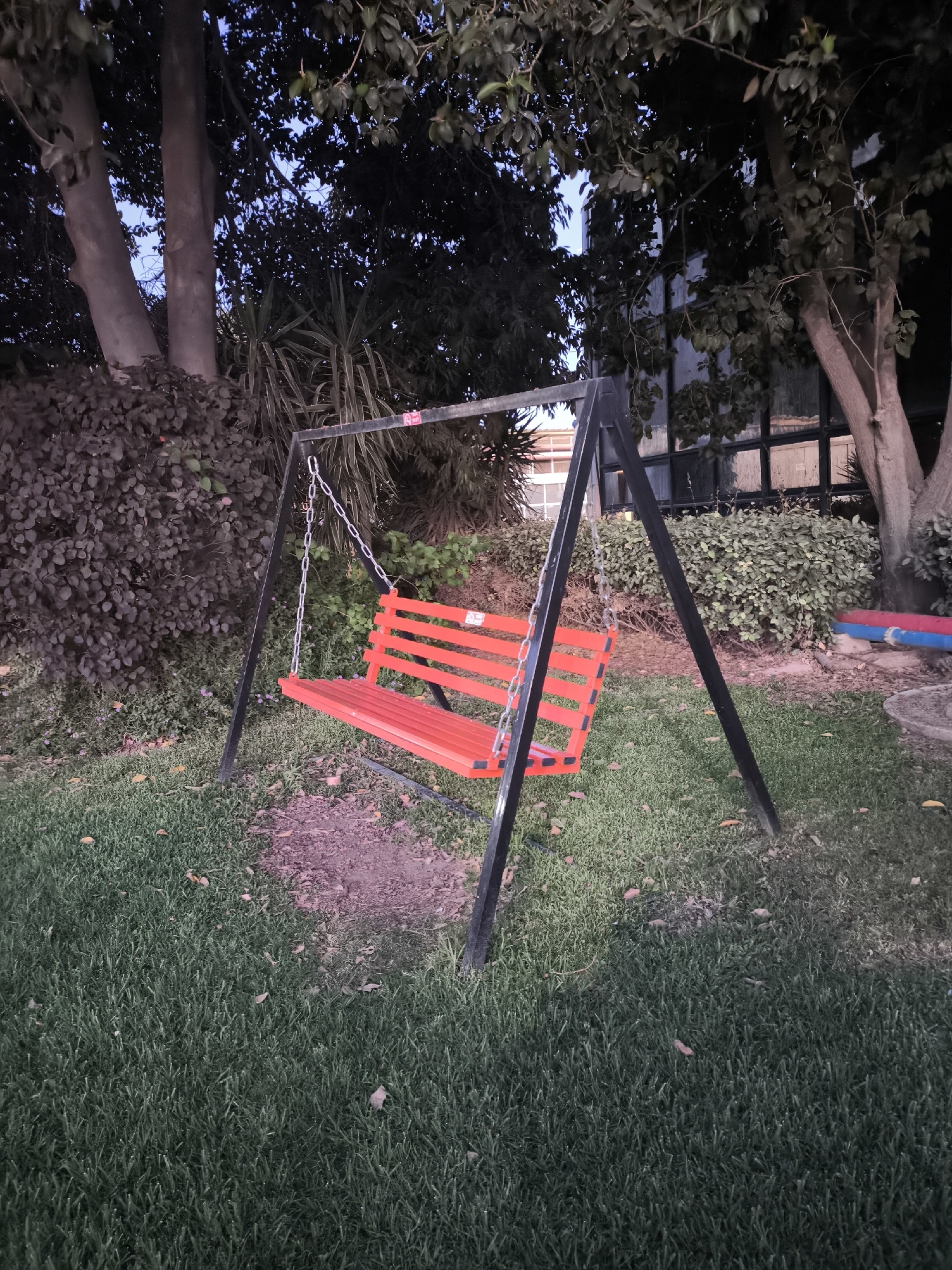 A red swing set is in the middle of a park or backyard with grass. A black frame supports the swing, which is also red with black chains. The background shows trees, bushes, and a building. It is nighttime, and there is some artificial light present.