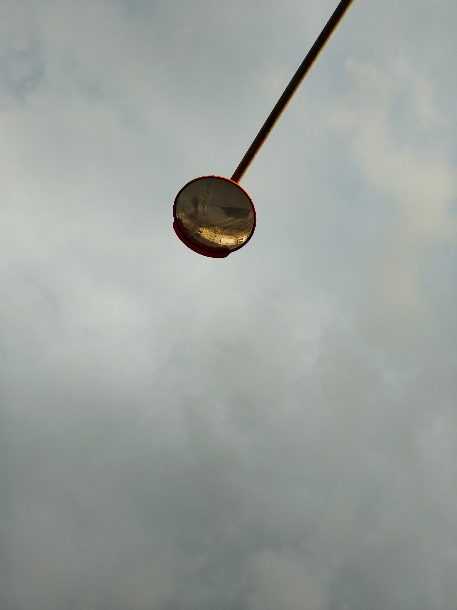 A low-angle shot captures a convex mirror attached to a diagonal pole, extending from the upper right towards the center of the frame, set against a cloudy sky. The pole is dark, possibly metal, and the mirror has a distinct red frame.
The reflective surface of the mirror shows a distorted image of a distant scene. Within this reflection, elements resembling buildings with windows and a stretch of ground or a road are visible, illuminated by a warm, golden light. This lighting suggests either early morning or late afternoon sun.
The sky is predominantly light gray, indicating an overcast or partly cloudy day, with some areas appearing darker and others slightly lighter due to varying cloud densities. No specific activities or events are depicted, and no text is visible. The mirror's position and type suggest its use for enhancing visibility around blind spots, commonly found on roads, at intersections, or in parking areas.