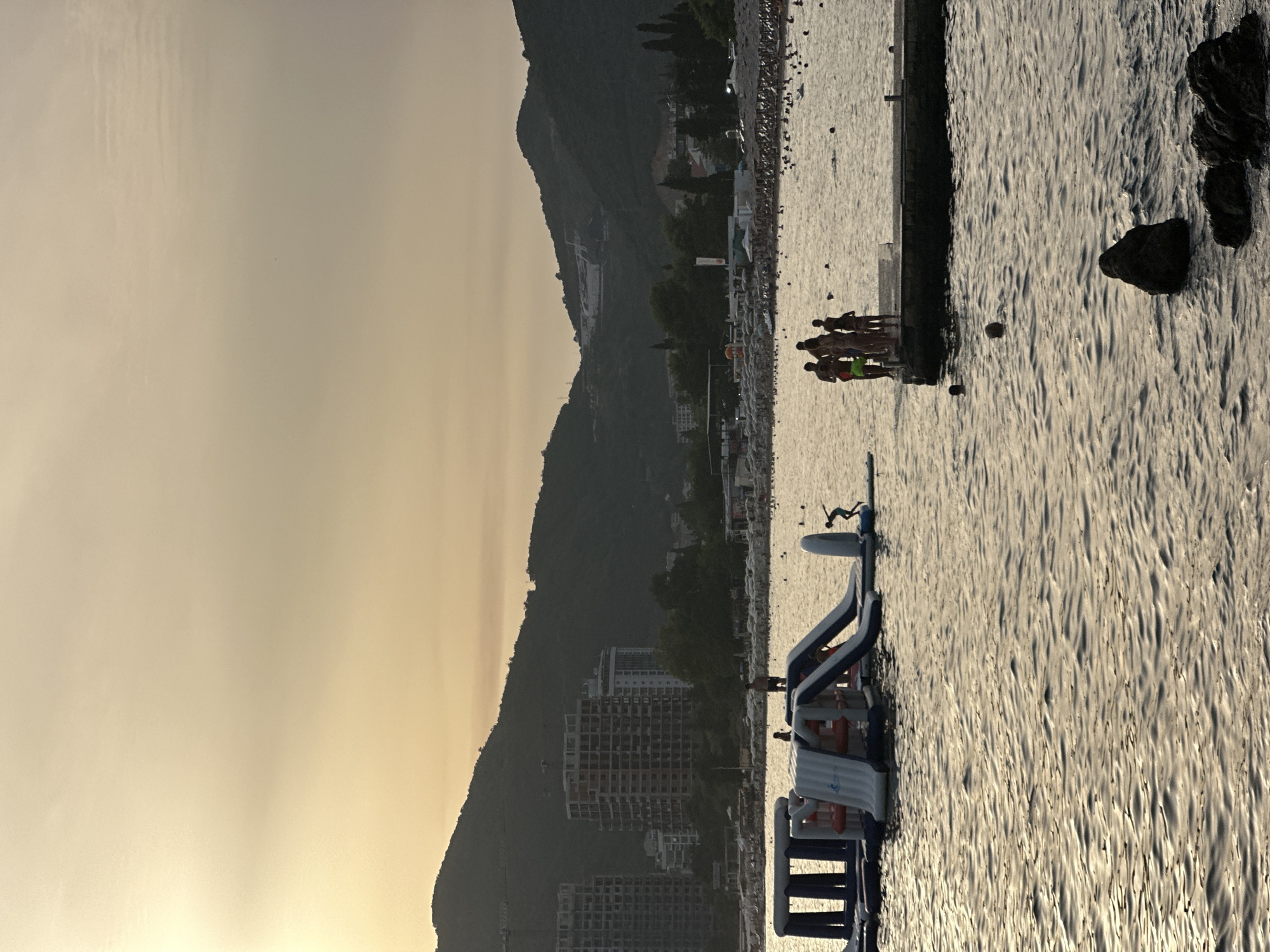 This image depicts a beach scene in Budva, Montenegro during a sunset. In the foreground, the water is rippled with golden light reflecting off the surface. A group of people is standing on a pier to the right, with a few individuals in the water around it. Further in the distance, an inflatable water park with slides is visible. Along the coastline, a densely populated beach is lined with umbrellas and trees. In the background, two unfinished high-rise buildings stand against a backdrop of dark, mountainous terrain. The sky is a soft, hazy yellow, indicating the approaching twilight.