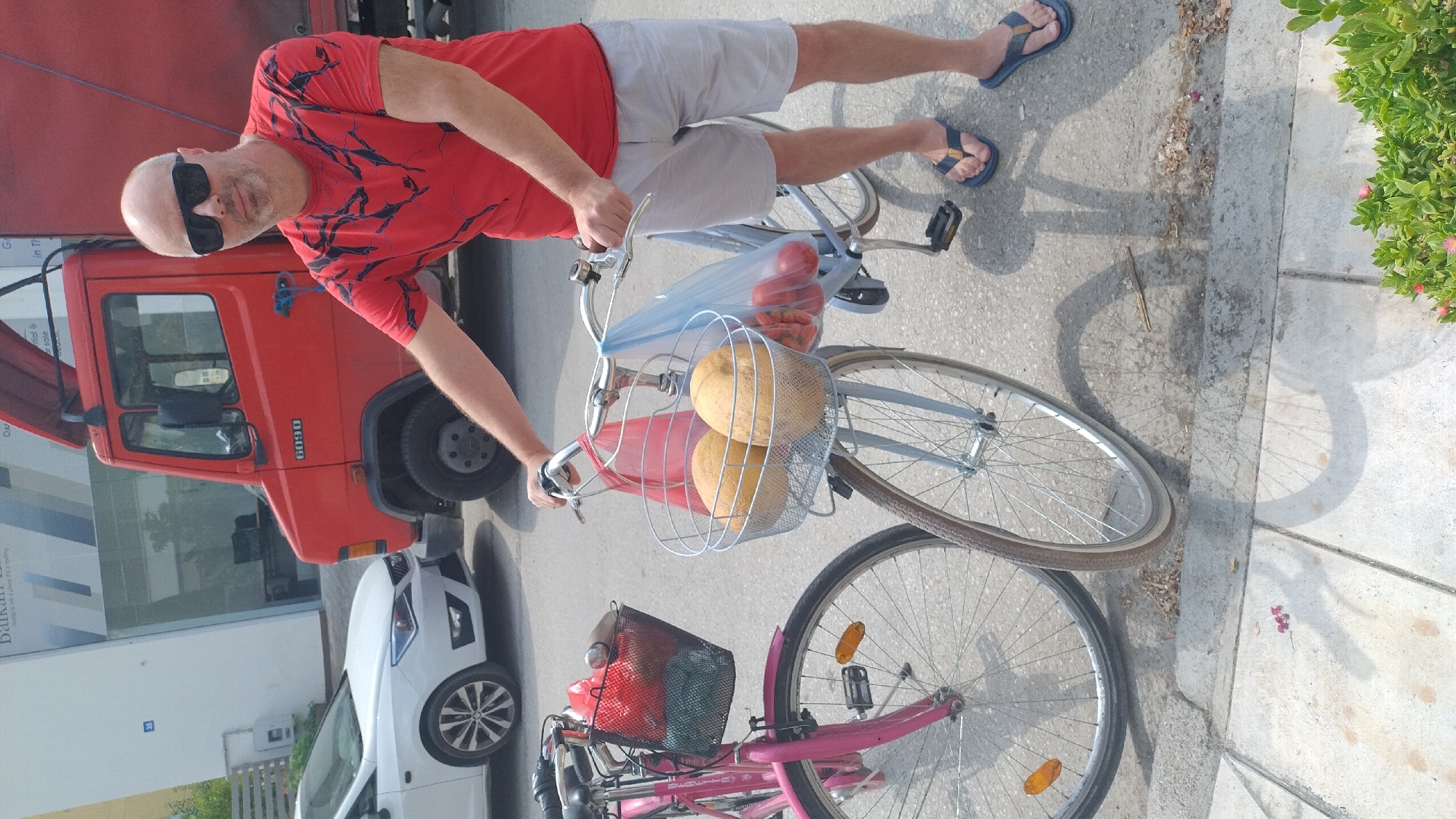 In Neoi Epivates, Greece, a bald man wearing sunglasses, a red t-shirt with a black pattern, and grey shorts stands beside two bicycles. One bicycle is pink and has a black basket containing red and green items. The other bicycle is light blue and has a silver basket with two large yellow fruits and a plastic bag filled with red tomatoes. A red truck is visible in the background, along with a white car parked in front of a building. The scene takes place on a paved street with concrete sidewalk.