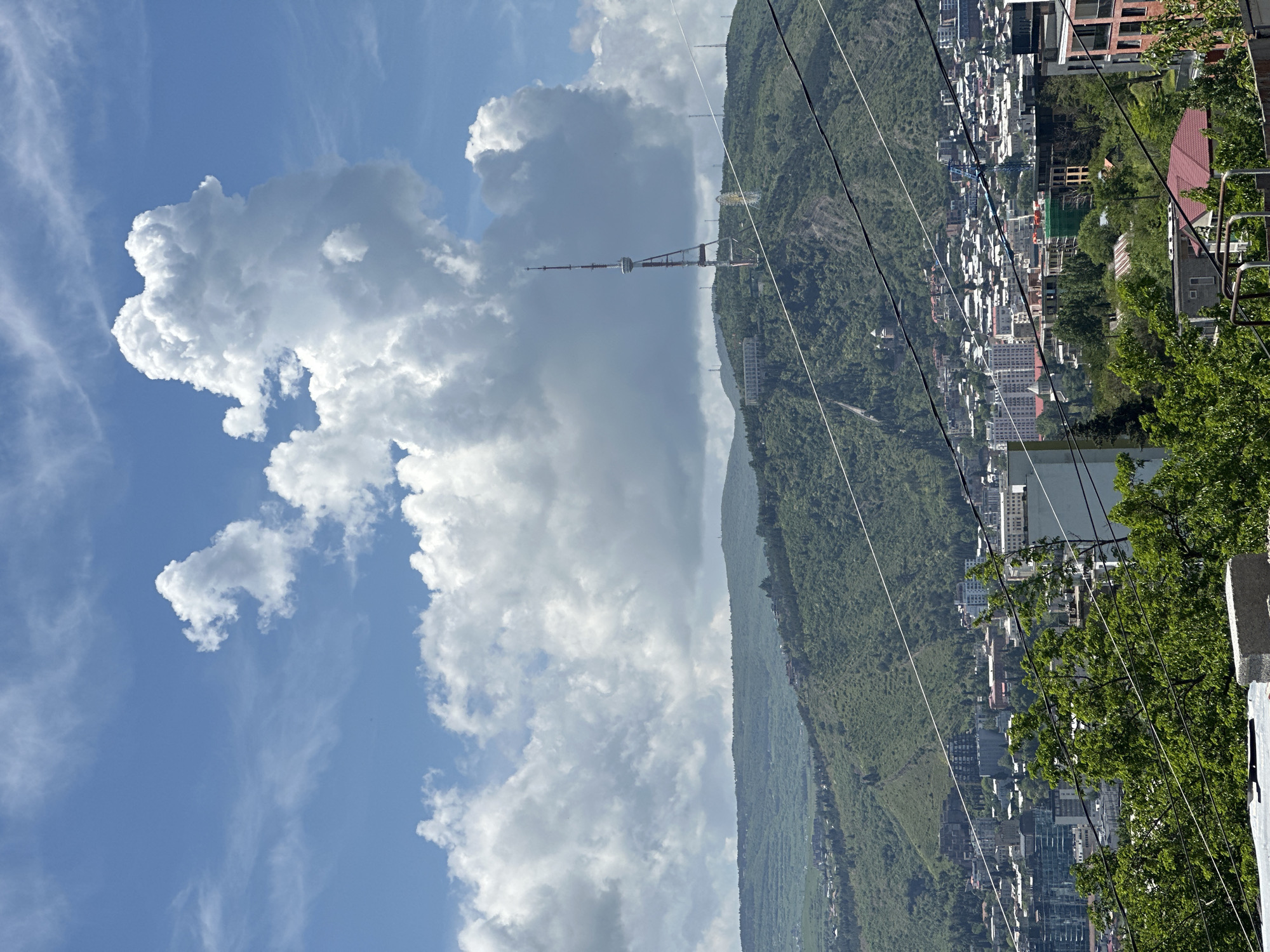 The image shows a cityscape of Tbilisi, Georgia, including the Tbilisi TV Broadcasting Tower. The view appears to be taken from an elevated position, possibly from a cable car or high vantage point overlooking the city. A lush green mountain partially obscures a view of the cityscape. In the sky, there are scattered cumulus clouds. The scene is captured in daylight, suggesting fair weather. Power lines are visible in the right side of the frame.