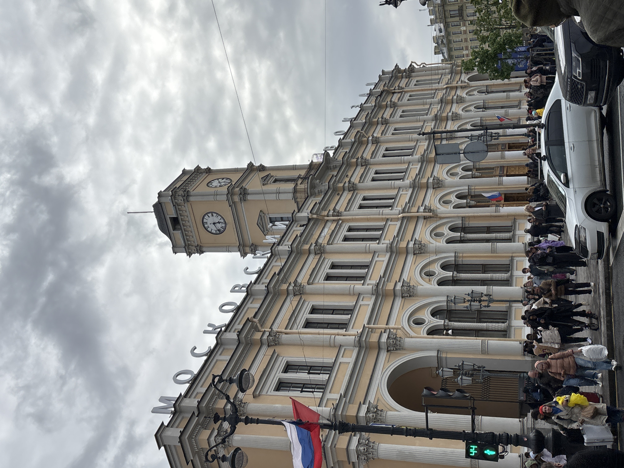 The image captures the Moskovsky Prospekt in Saint Petersburg, Russia, featuring a prominent architectural building with a clock tower and the Russian inscription "МОСКОВСКИЙ" (Moskovsky) along the facade. The building is light yellow with white accents. A Russian flag is attached to a lamppost in front of the building. A crowd of people is present on the sidewalk adjacent to the building. Two vehicles, one white and one dark colored, are parked on the street. The sky is overcast with a gray, cloudy appearance. A traffic light is visible, indicating green, which may indicate that pedestrians are authorized to cross. The overall setting is a street scene, with a focus on the architecture and the people gathered near the building.