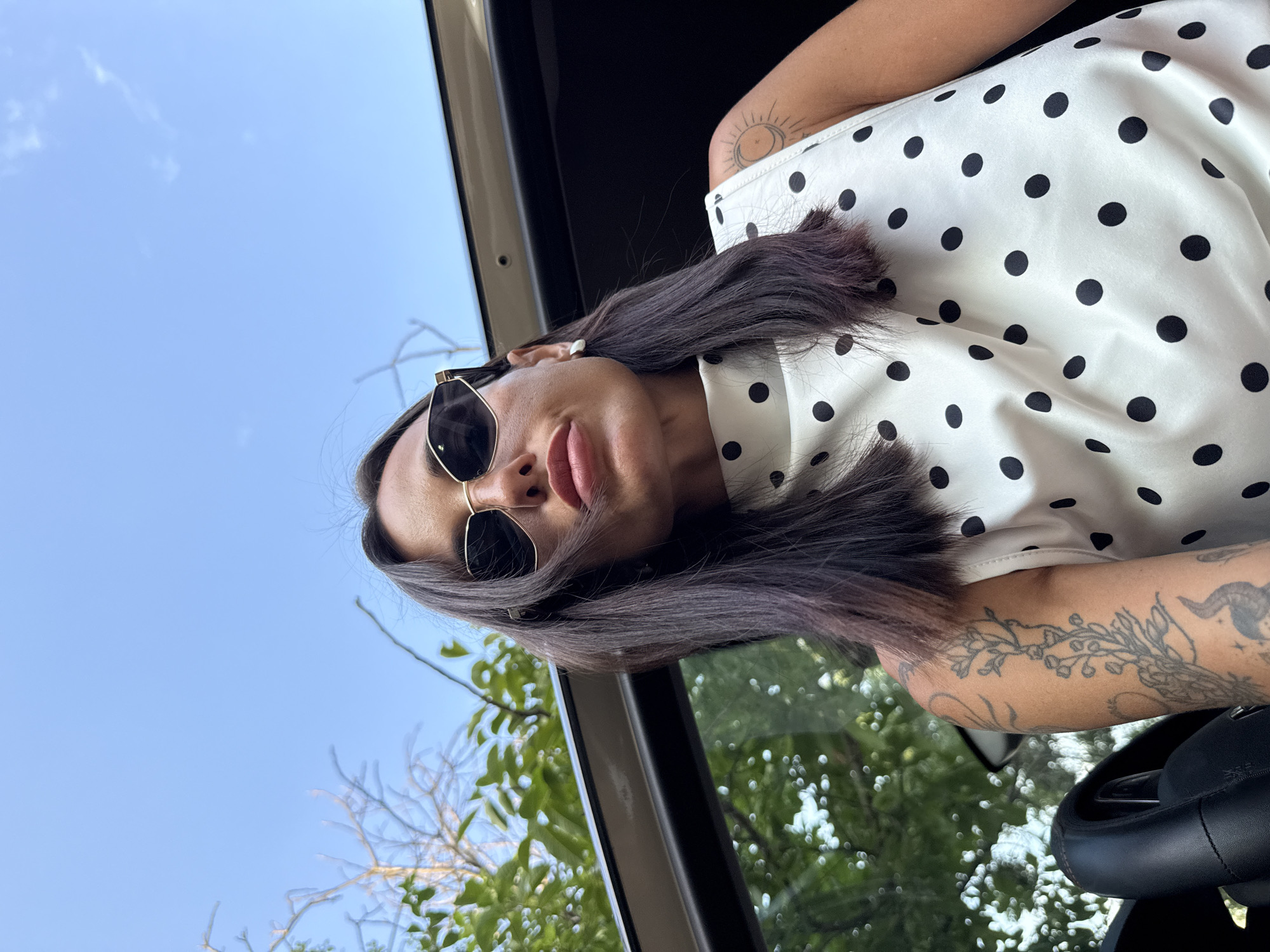A woman with dark hair and sunglasses is taking a selfie from a low angle. She is wearing a white sleeveless top with black polka dots and has tattoos on her arms. She is sitting in a car, with the sky and trees visible in the background. The location is not specified in the image.