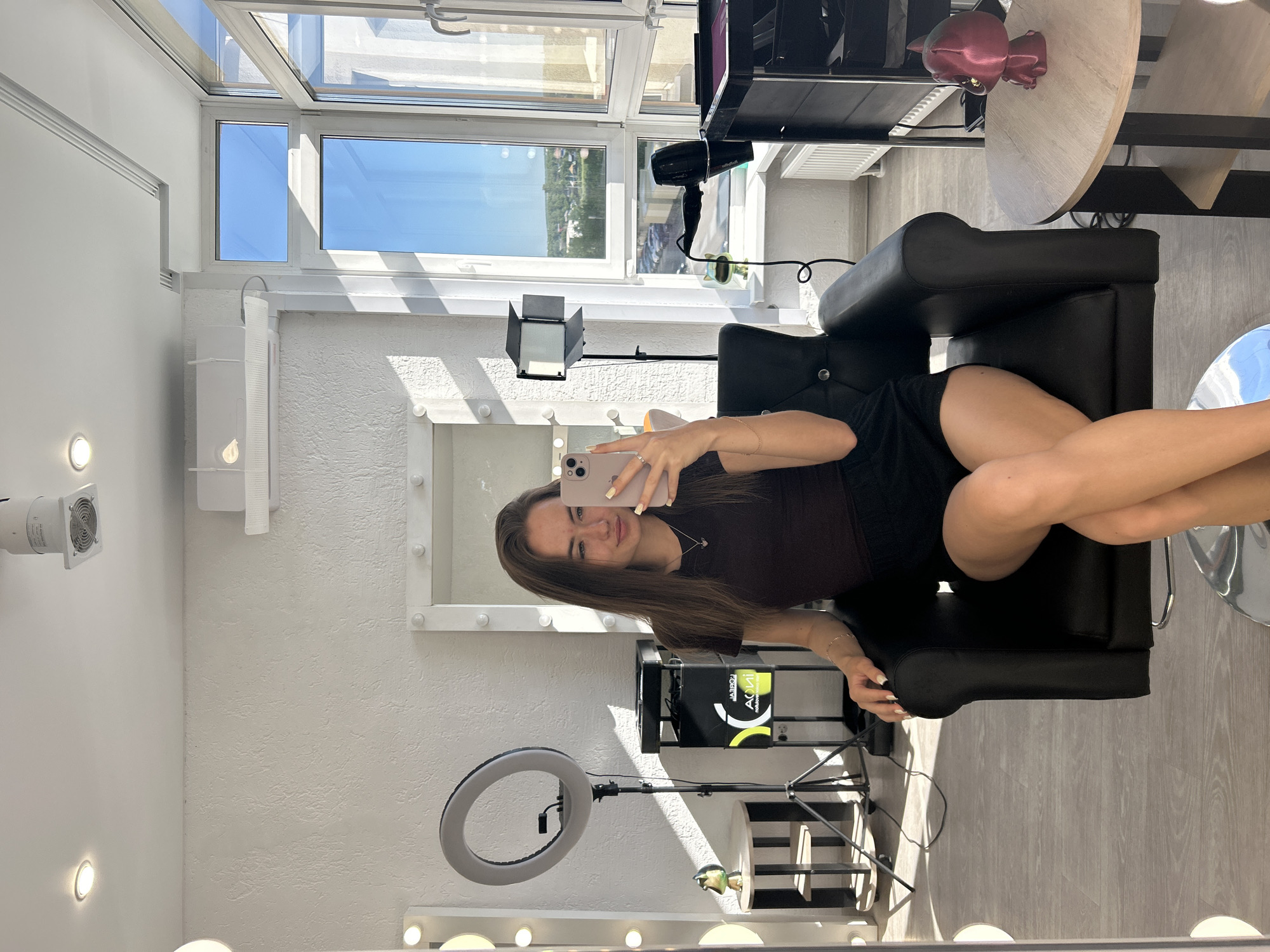 A young woman is seated in a black leather salon chair, holding a pink smartphone and taking a selfie. She is wearing a dark brown t-shirt and black shorts. Her legs are crossed, and her right arm is resting on the armrest of the chair. The background appears to be a brightly lit salon or studio, with large windows offering a view of a suburban landscape. Several pieces of equipment are visible, including a ring light on a tripod, a large mirror with lights around its frame, a hair dryer, and a rolling cart with supplies. A small, dark red statuette sits on a round wooden table to the right of the woman. The overall impression is one of a professional or personal beauty space.