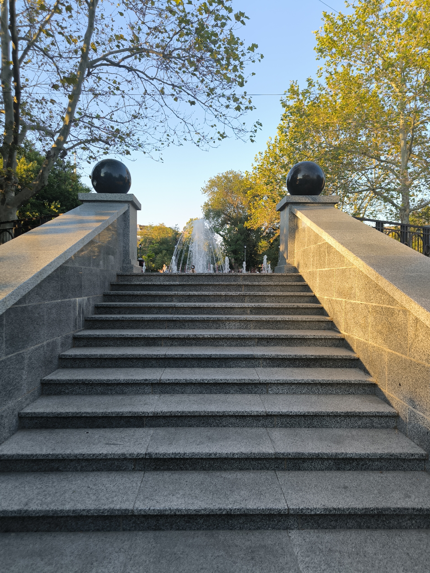 A wide, symmetrical stone staircase leads upwards towards a fountain in a park on a sunny day. The staircase is flanked by granite walls topped with large, dark spherical ornaments and balustrades. Trees with green foliage are visible in the background, framing the fountain which is spraying water into the air. A clear blue sky is overhead. The scene appears to be located in a public park, possibly in Mykolaiv, Ukraine, given the context provided. No people are clearly discernible in the immediate vicinity of the fountain, though there may be indistinct figures further in the distance. The overall impression is one of a peaceful and pleasant outdoor setting.