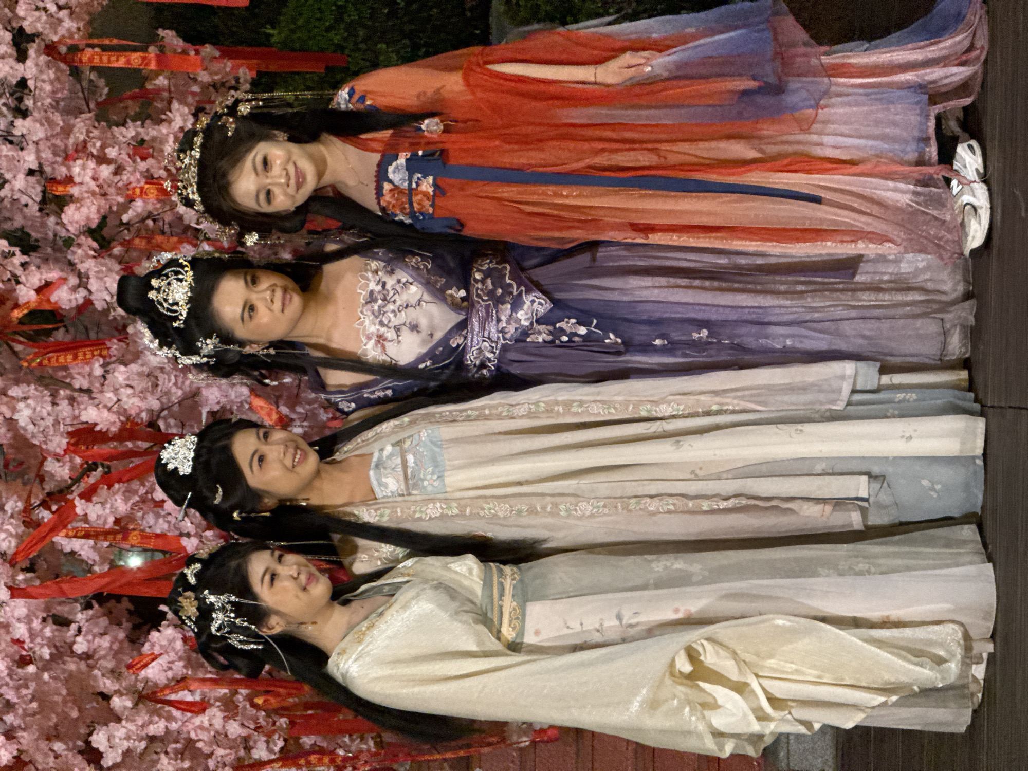 Four women wearing traditional Chinese dresses and ornate hairpieces are standing together. They are positioned in front of a blooming cherry blossom tree with red ribbons hanging from its branches. The dresses are different colors: orange, purple, and two shades of beige. The location is likely a park or outdoor setting in Jiulongpo District, China, given the presence of the cherry blossom tree and traditional attire.