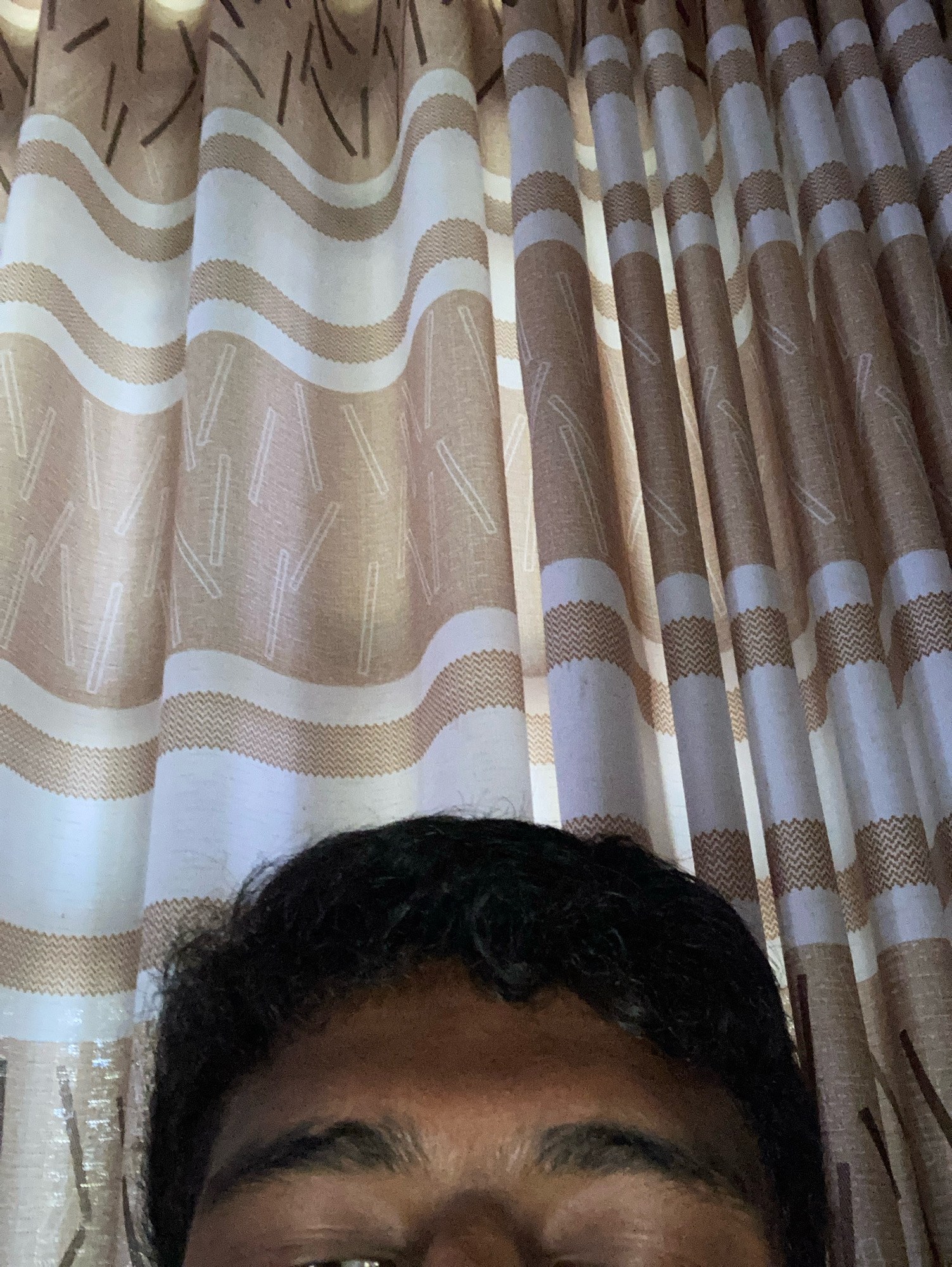The image shows a close-up view of the upper forehead and eyes of a person, with a patterned curtain in the background. The curtain features wavy horizontal bands of white and light brown, with a textured zigzag pattern within some of the brown bands. The brown bands are also adorned with scattered vertical and angled lines. The person's dark, curly hair is visible at the top of the frame, framing their forehead and eyebrows. The lighting suggests an indoor setting. There are no discernible landmarks or contextual elements within the image that would allow for the identification of a specific location in Daladagama, Sri Lanka.