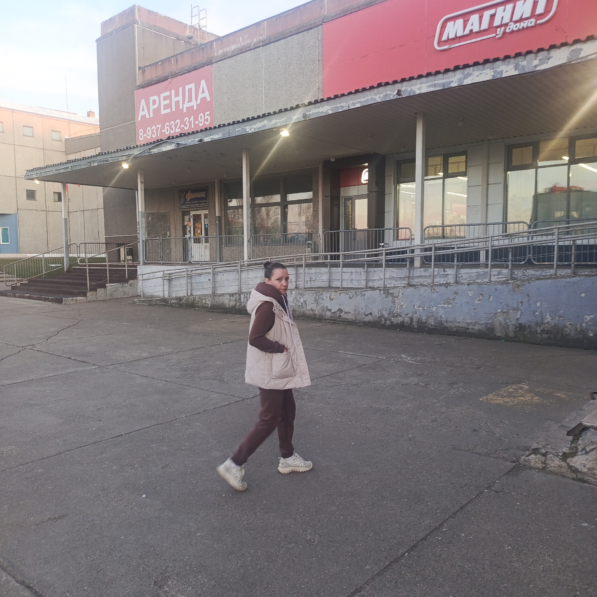 The image depicts a woman walking in front of a single-story building in Марковский, Russia. The building appears to house commercial establishments, indicated by signs for "Аренда" (Rent) and "Магнит у дома" (Magnet near the house), a retail chain. The woman is dressed in a brown tracksuit and a beige vest and is walking towards the left side of the frame. The building has a dated facade with a covered walkway supported by pillars. A railing is visible, possibly for accessibility. The area in front of the building is paved with concrete. The overall impression is that of a typical urban or suburban landscape in Russia.