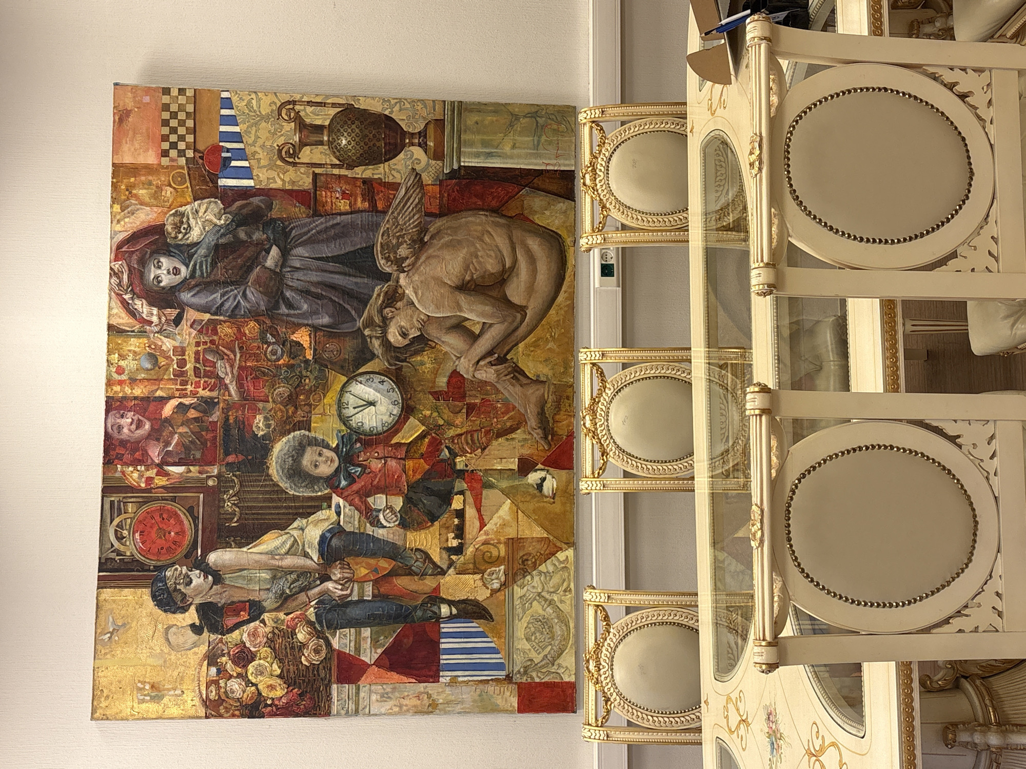 The image shows a section of a wall where an art piece hangs. The artwork is a collage-like painting that includes figures such as a woman holding a small dog, a nude angel, two people with a basket of flowers, and other imagery like clocks and various patterns. Adjacent to the artwork is a cream-colored cabinet with glass panels and ornate gold trim, featuring oval backings visible inside the cabinet. The wall and cabinet appear to be inside a building, possibly a home or a gallery. There is no information in the provided image to determine the specific location in Moscow, Russia.