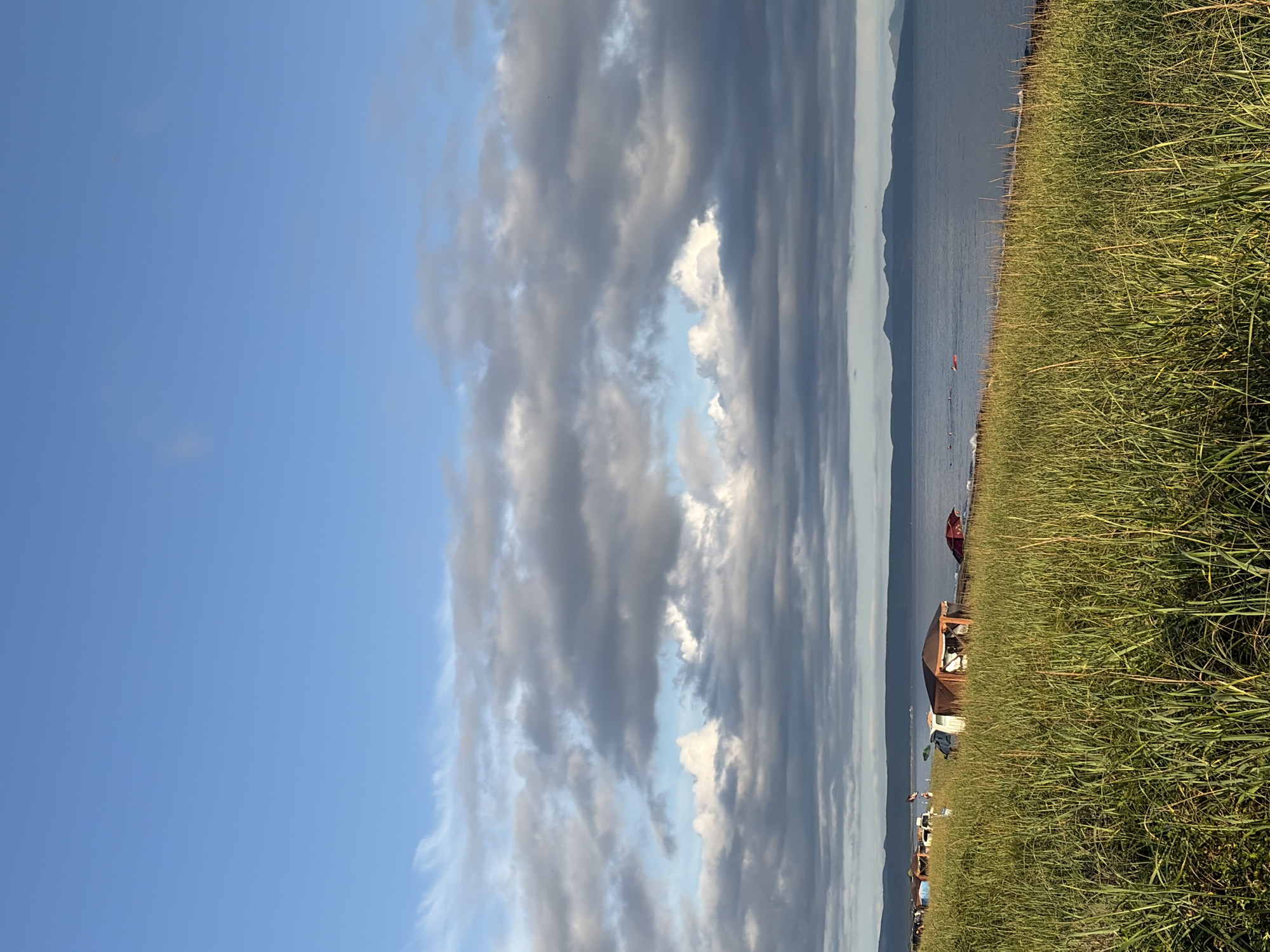 The image depicts a coastal scene with a foreground of tall, green grass sloping down towards a body of water. Several small structures, resembling tents or shelters, are visible along the shoreline, with some vehicles parked nearby. In the distance, a range of mountains forms the horizon, with a calm sea between the shore and the mountains. The sky above is a mix of blue and dramatic grey clouds. The image appears to be taken in the city of Ozerskoe, Russia, given the context. There are no clear indications of specific people or their actions. The overall environment suggests a natural, possibly recreational area.