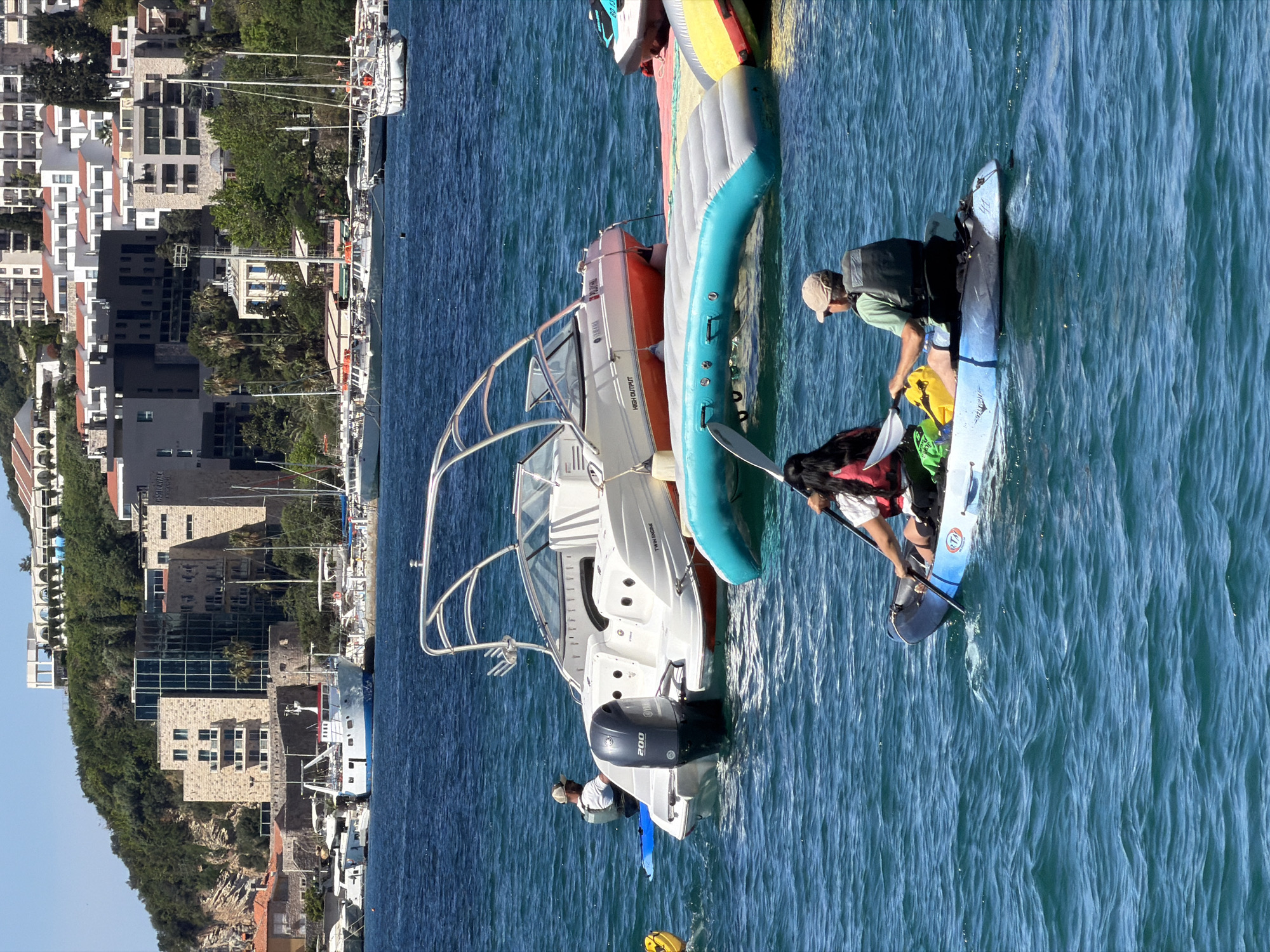 A partly cloudy, sunlit exterior shot captures a marine scene in Budva, Montenegro. A white and orange Yamaha motorboat is anchored in turquoise waters, with two people on board. One person stands by the engine, and another appears to be helping someone onto a blue paddleboard from an inflatable multi-person raft.
The coastline of Budva is visible in the background, featuring various buildings and structures, including a hotel and docked sailboats. The water's surface displays a textured pattern of ripples.