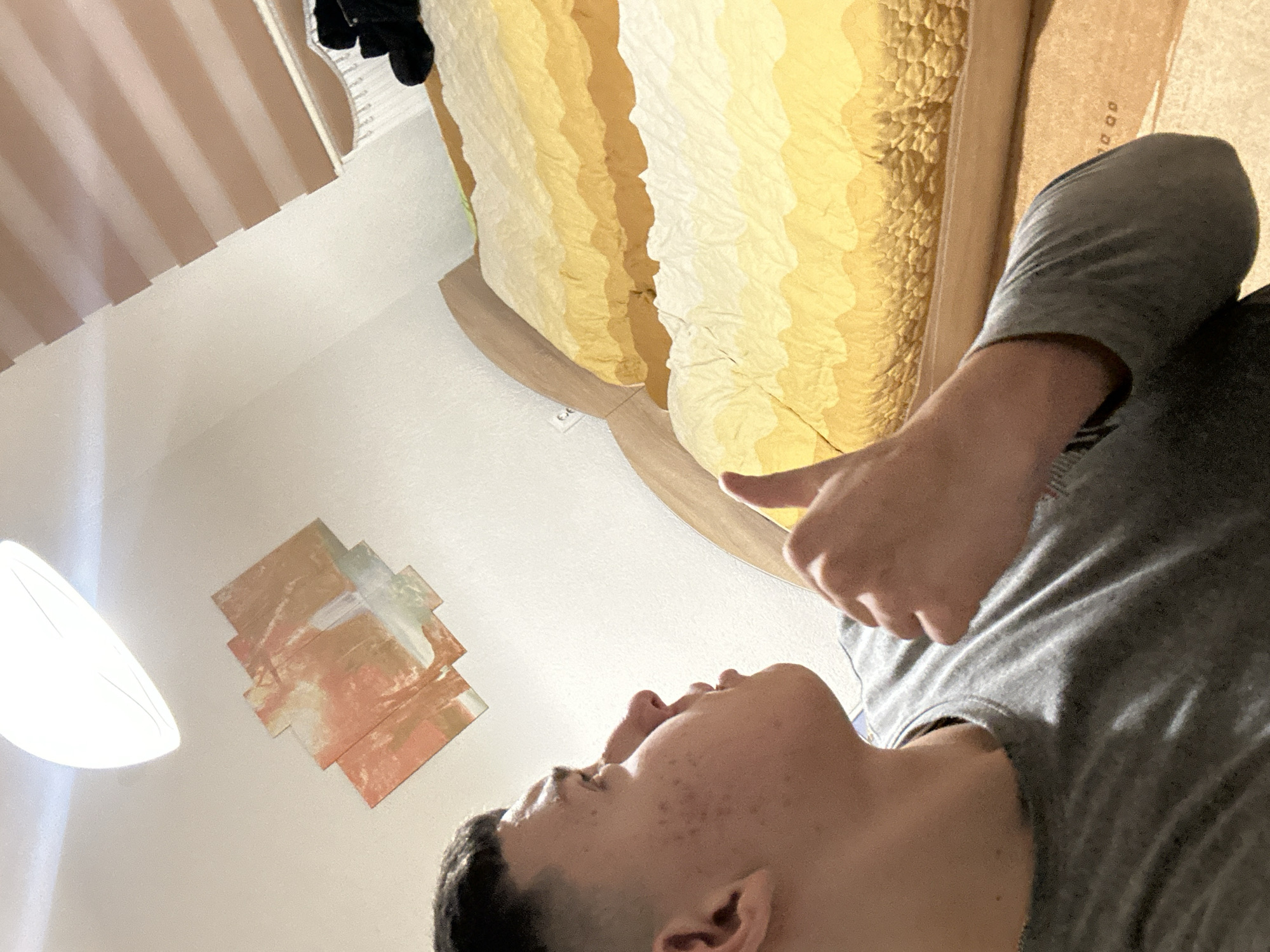 This image depicts an indoor scene in Zlatograd, Bulgaria, observed from a low angle. A male individual, visible from the chest up, occupies the left foreground. He has short, dark hair with faded sides, and blemishes are present on his right cheek and jawline. He is wearing a grey, long-sleeved crew-neck shirt and looks upwards and slightly to the right with his mouth slightly open. His right hand is raised, performing a "thumbs up" gesture.
To the right, a bed with a light brown, arched wooden headboard is present. The bed is covered with a comforter or duvet featuring horizontal bands of color ranging from pale yellow to darker gold and brown. A dark, possibly black, object is situated at the foot of the bed in the upper right corner.
The walls are light-colored. A luminous white, oval-shaped ceiling light fixture is centered at the top of the frame. Below the fixture, four rectangular abstract art pieces, featuring shades of orange, brown, and white, are mounted on the wall, appearing slightly offset. In the upper right background, a window is fitted with a horizontally striped blind, likely in shades of brown or beige. A white double electrical outlet is visible on the wall behind the headboard of the bed. The floor, glimpsed at the bottom right, appears to be a light-colored, textured surface.