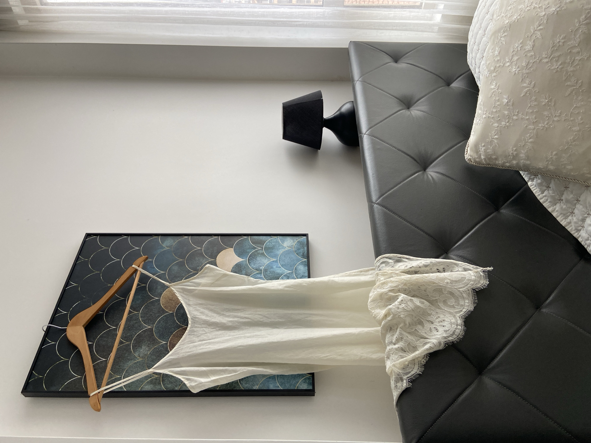 A low-angle shot reveals a room interior. The room features a black tufted bench against a white wall. The corner of a window is visible at the top. A black sconce is mounted on the wall above the bench. On the wall, a framed artwork with a teal and blue fish-scale pattern is mounted. A wooden hanger displays a white lace-trimmed top. A white decorative pillow sits on the bench. The location is not discernible from the image.