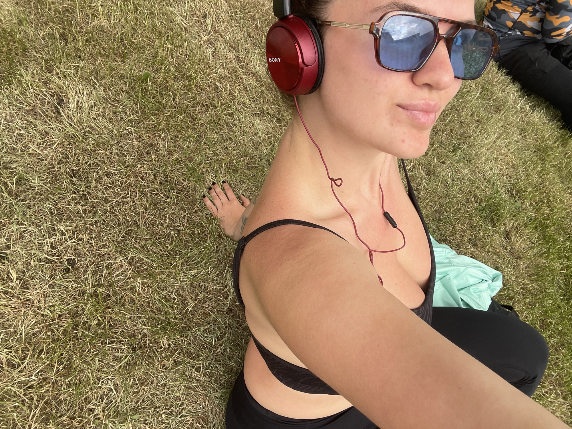 A woman is sitting on grass in a park. She is wearing a black sports bra and black leggings. She has red Sony headphones on and is wearing sunglasses. There is a light green shirt or towel behind her. A person is lying on the grass behind her to the right. The grass appears to be dry. It is not possible to determine the location as Moscow, Russia, based on the information provided in the image.