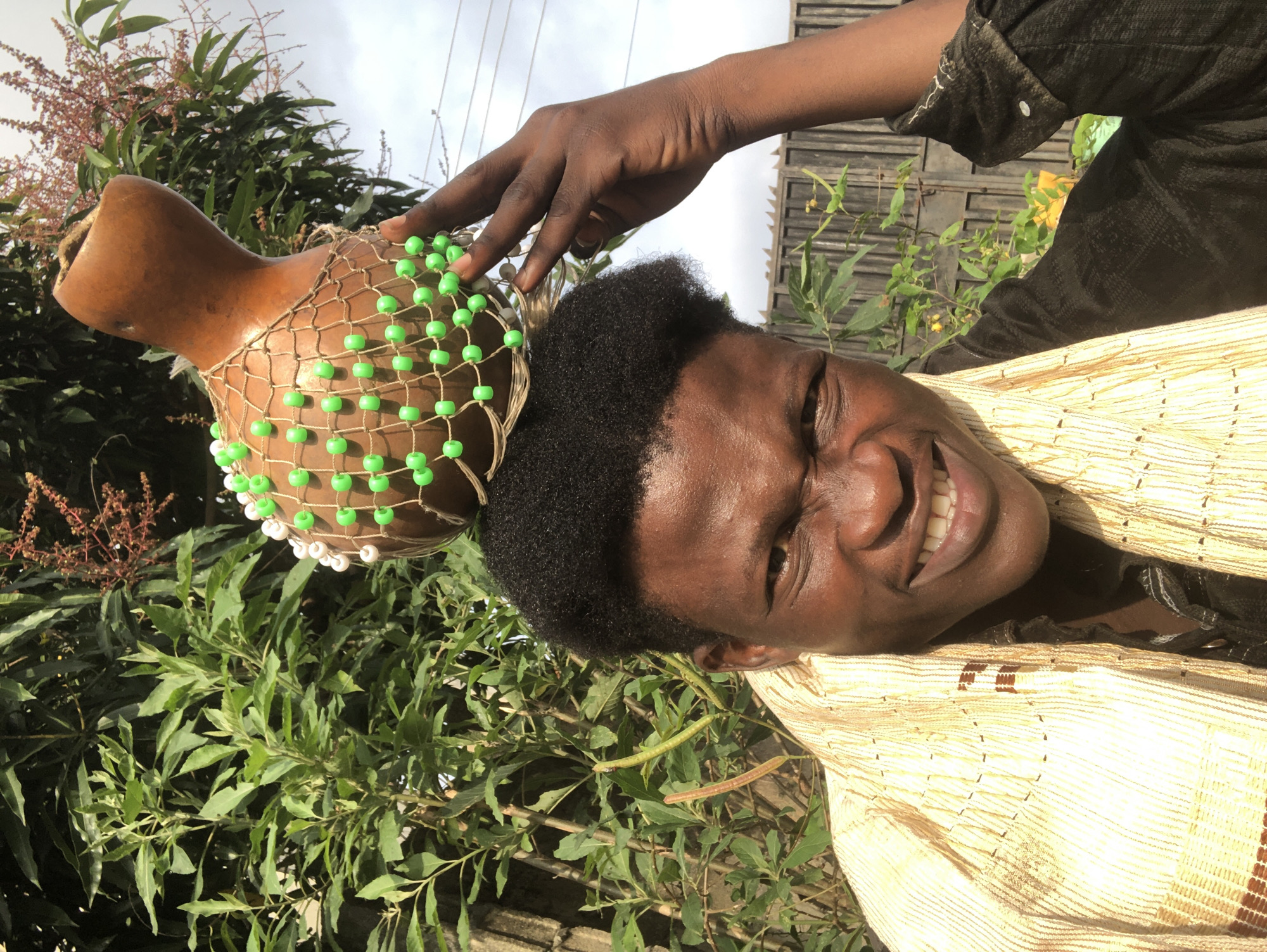 A close-up, medium shot captures a young Black male from the chest up, outdoors in Ado, Nigeria, with a calabash decorated with green and white beads resting on his head. He is wearing a patterned light-colored garment draped over his shoulders and a dark top underneath. The subject is smiling broadly, with his mouth slightly open, revealing his teeth. His right hand is visible, holding the calabash in place. Behind him, lush green foliage and a wooden fence create a natural backdrop. The lighting suggests it is daytime, possibly late morning or early afternoon, with sunlight casting shadows. The overall impression is one of warmth and vibrancy.