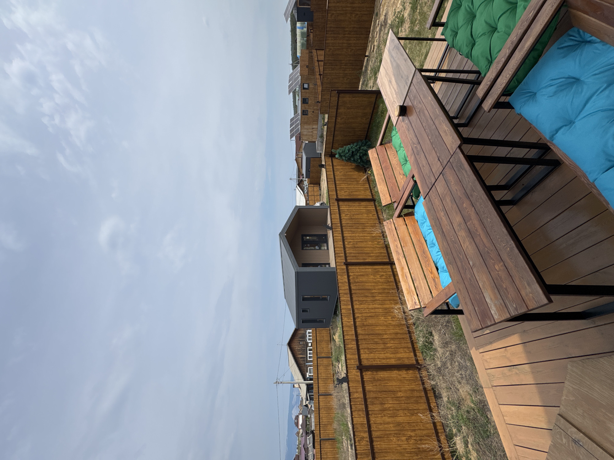 A wide-angle, high-angle outdoor shot captures a wooden deck with a dining table and benches in the foreground, looking towards a residential area in Khuzhir, Russia. The deck is furnished with two wooden benches, one with blue cushions and the other with green cushions, and a large wooden dining table with a light-colored tabletop and dark metal legs. A small ashtray sits on the table. Beyond the deck, a brown wooden fence encloses a grassy yard. In the background, several modern, modular houses are visible, some with dark gray siding and metal roofs. One house features a light brown exterior and a dark roof. A small, artificial Christmas tree stands to the left of the frame. Solar panels are mounted on the roofs of some of the buildings. The sky is overcast with light gray clouds. In the distance, faint mountains can be seen. The scene depicts an outdoor recreational area in a suburban setting.